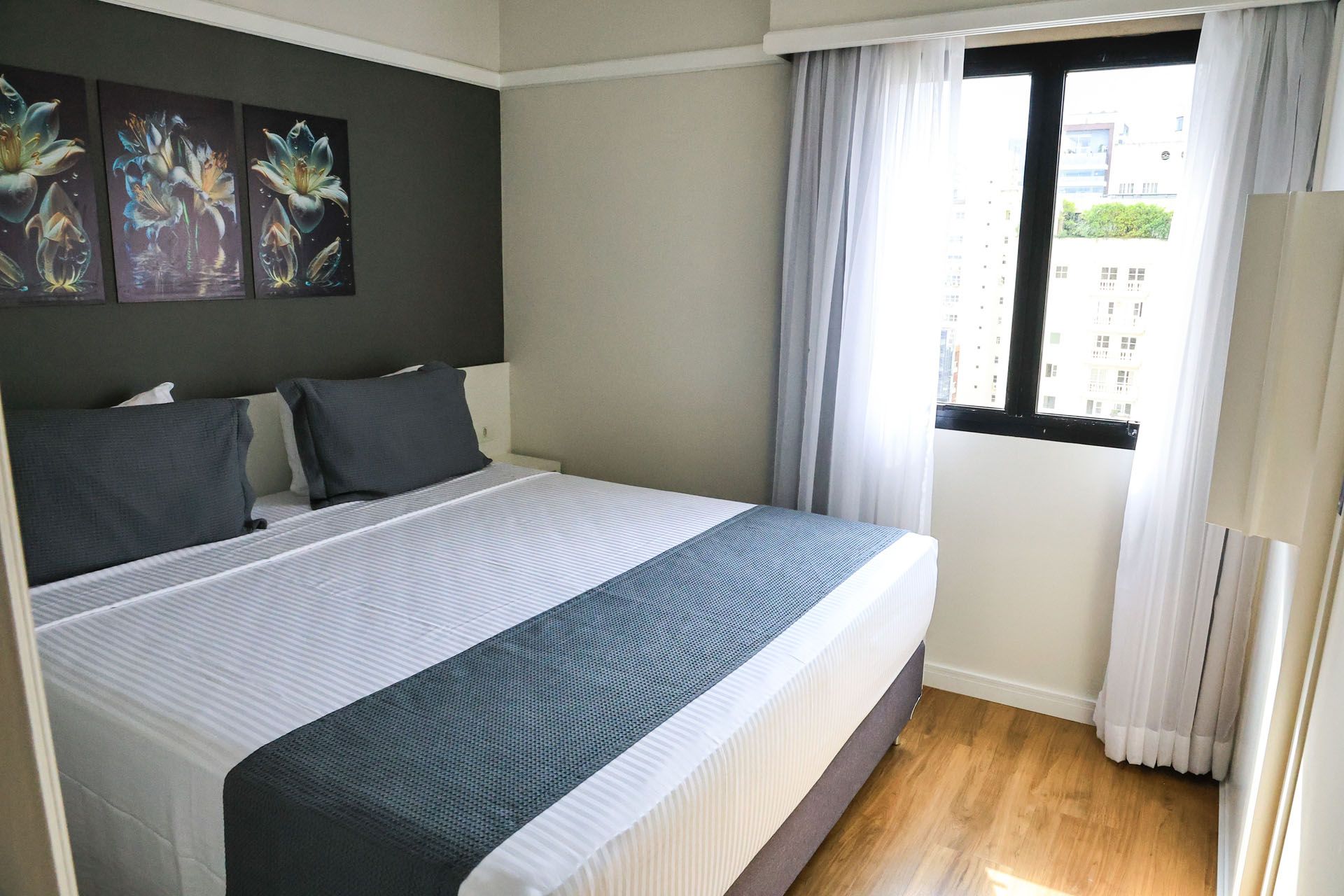 Quarto iluminado com cama arrumada, travesseiros cinza-escuros, obras de arte na parede e janela com cortinas brancas.