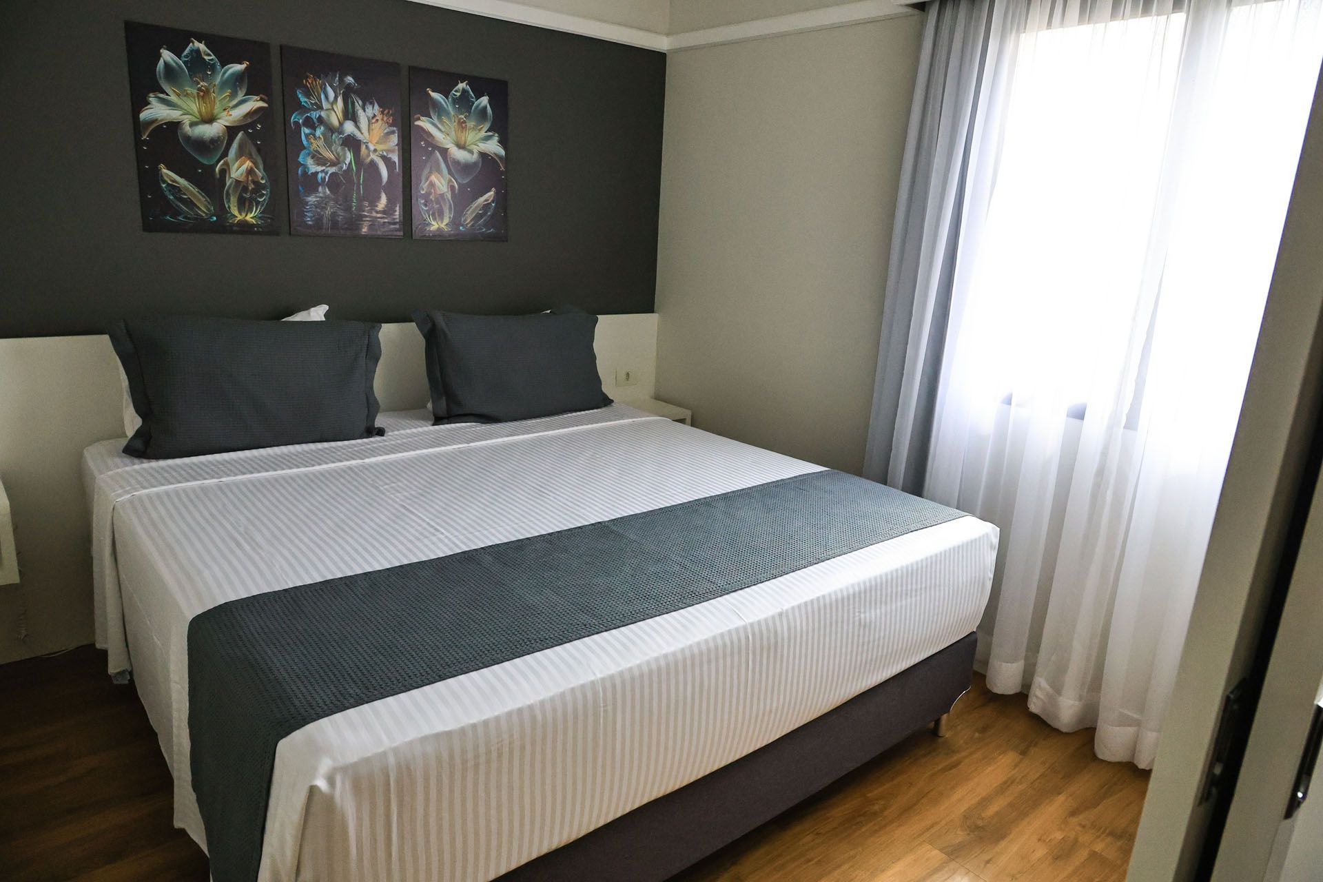 Quarto moderno com cama arrumada, travesseiros escuros, obras de arte na parede e cortinas translúcidas junto a uma janela bem iluminada.