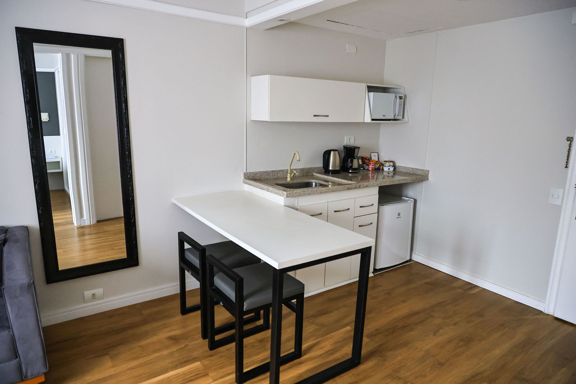 Cozinha pequena com armários brancos, cadeiras pretas e uma mesa ao lado de um espelho de parede.
