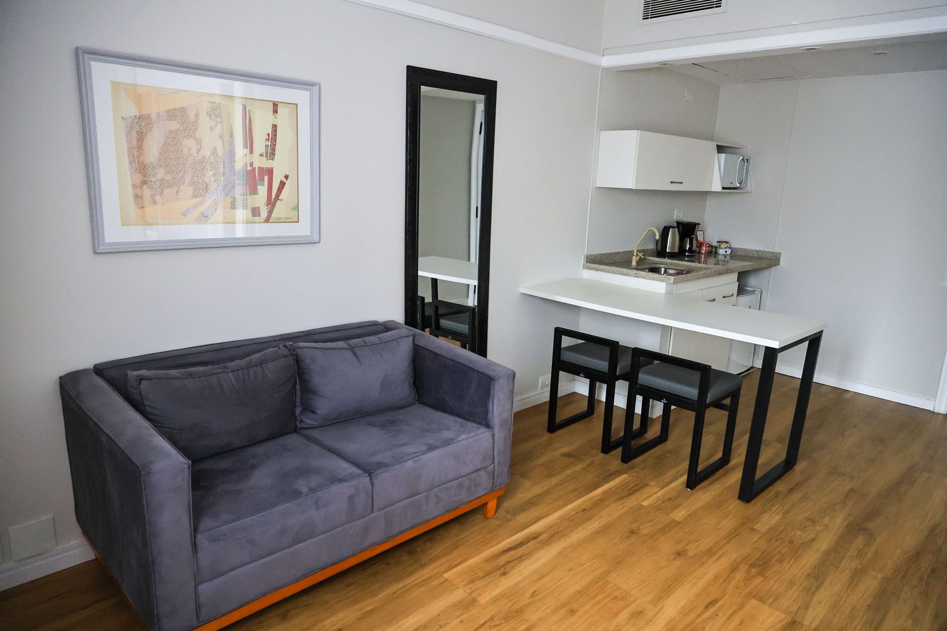 Sala de estar moderna com sofá cinza, obras de arte na parede, espelho e pequena cozinha americana sobre piso de madeira.