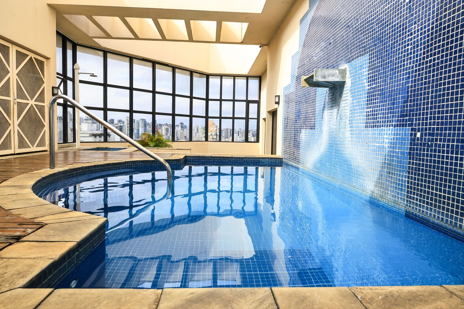 Piscina interna com azulejos azuis, parede de vidro curvada e cascata.