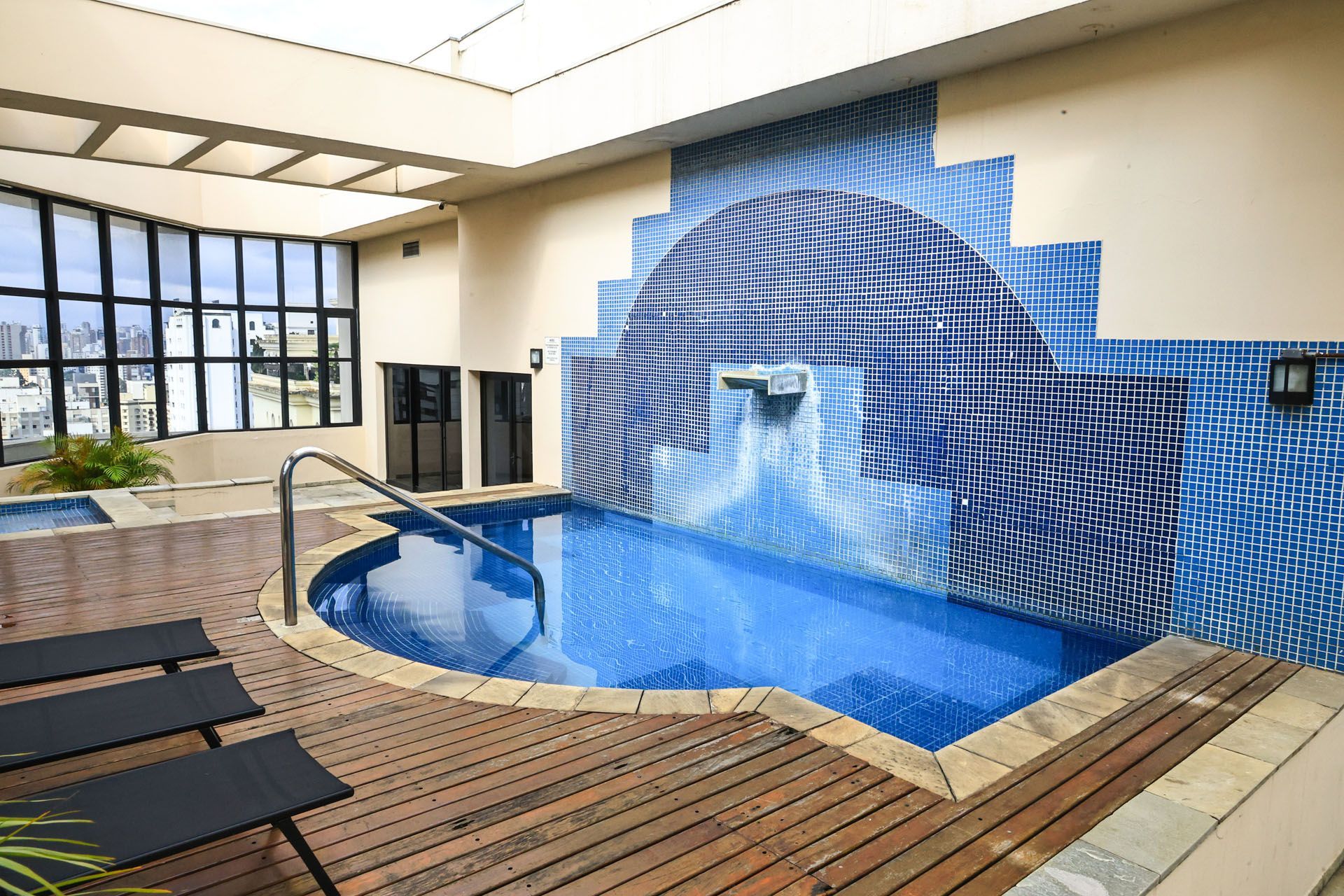 Piscina de spa interna com cascata de mosaico azul e área de estar com deck de madeira.