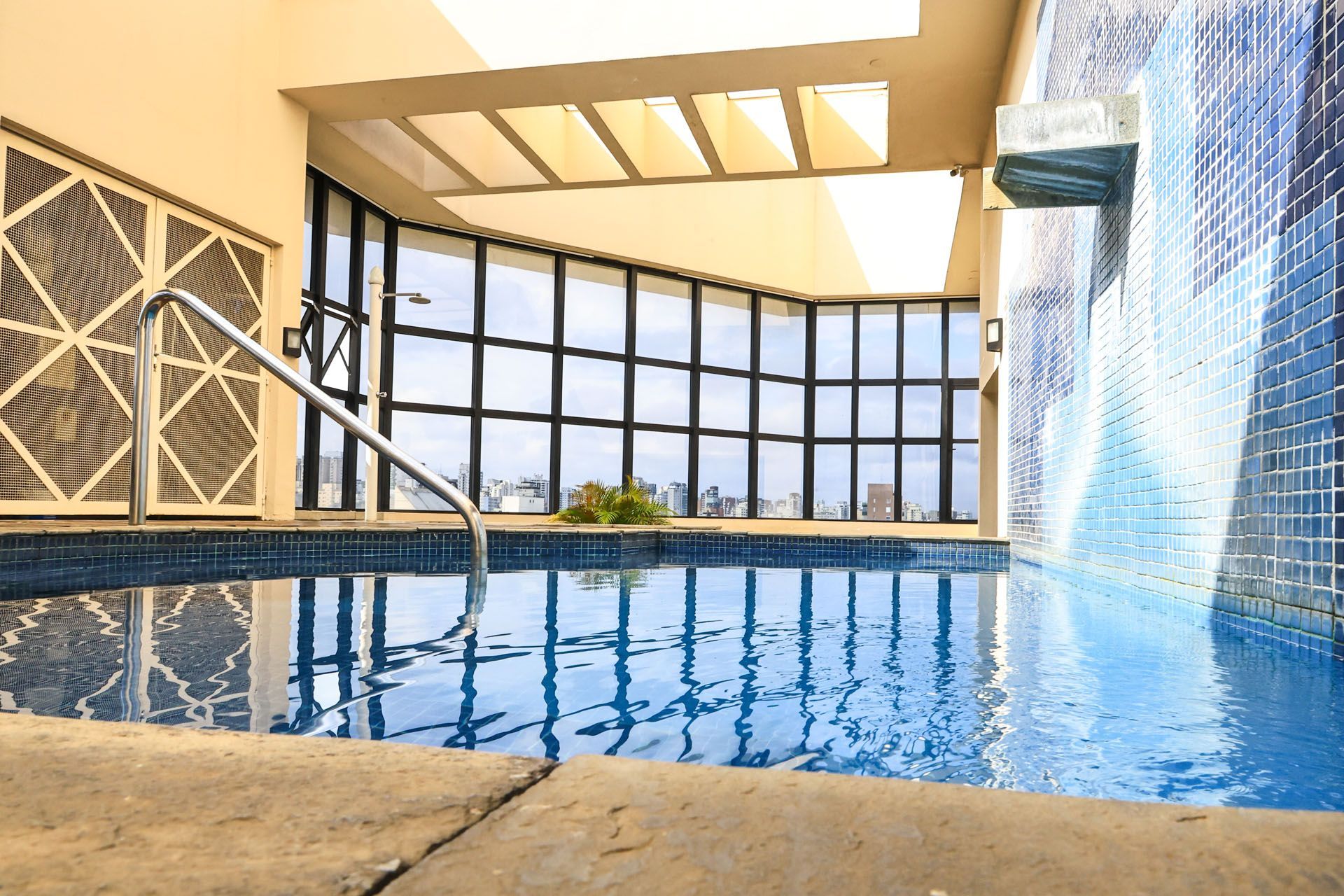 Piscina coberta com água azul, paredes revestidas de azulejos e grandes janelas com vista para a cidade.
