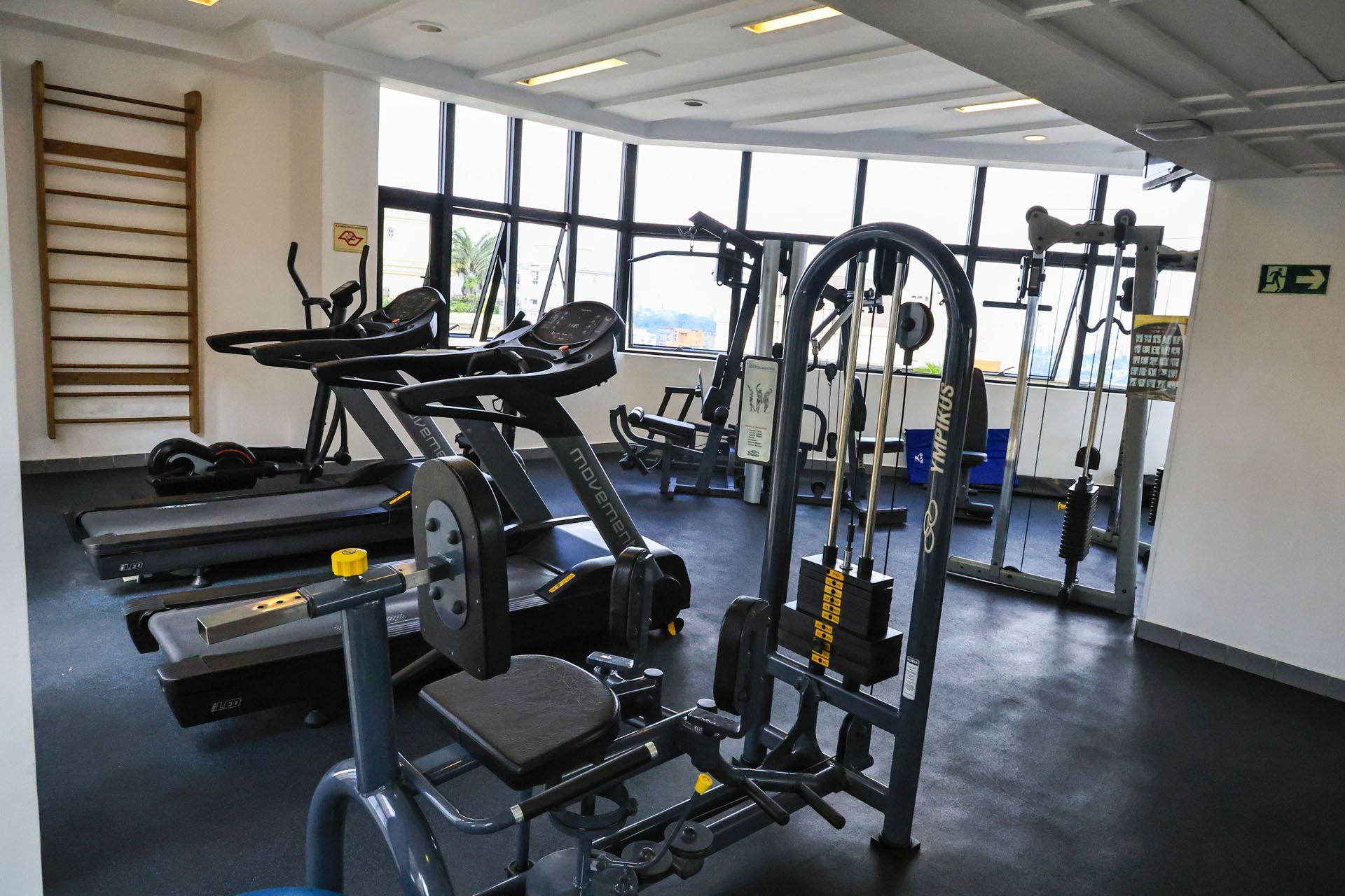 Sala de ginástica vazia com esteiras, máquinas de musculação e equipamentos de ginástica perto de grandes janelas.