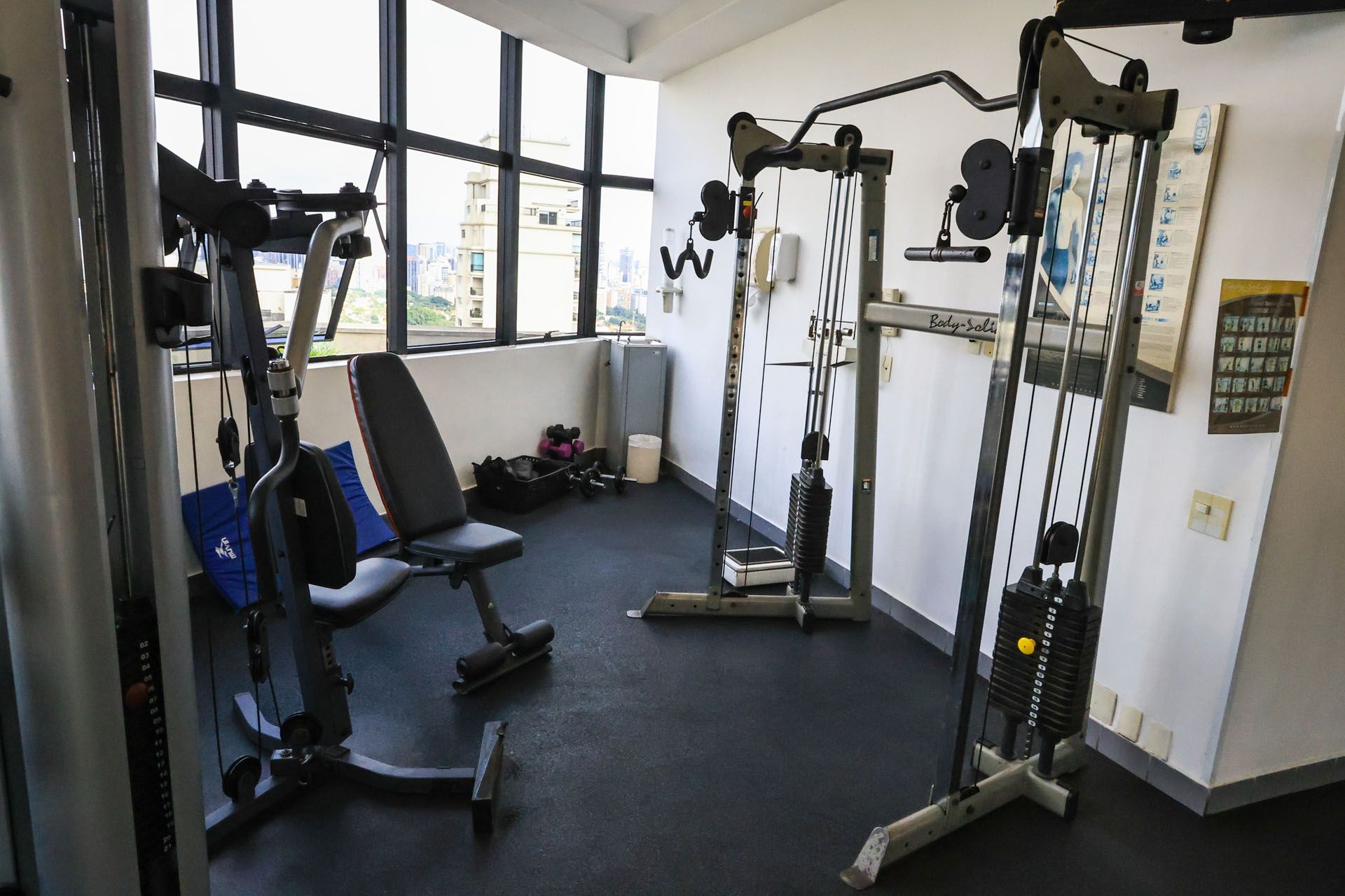Pequena sala de ginástica com aparelhos de musculação, estação de cabos para pesos e banco perto das janelas.