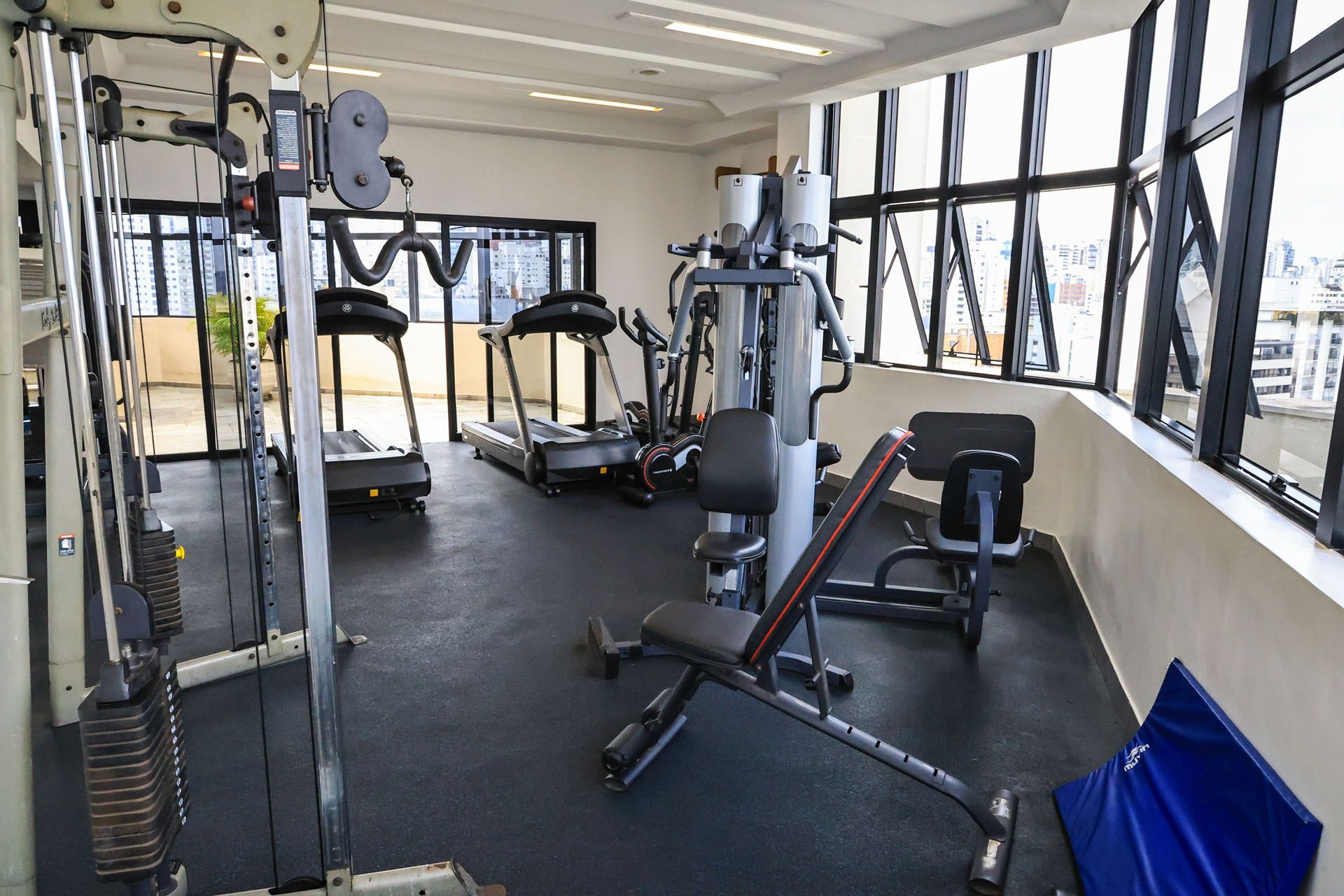 Pequena sala de ginástica com esteiras, aparelhos de musculação e grandes janelas com vista para o exterior.