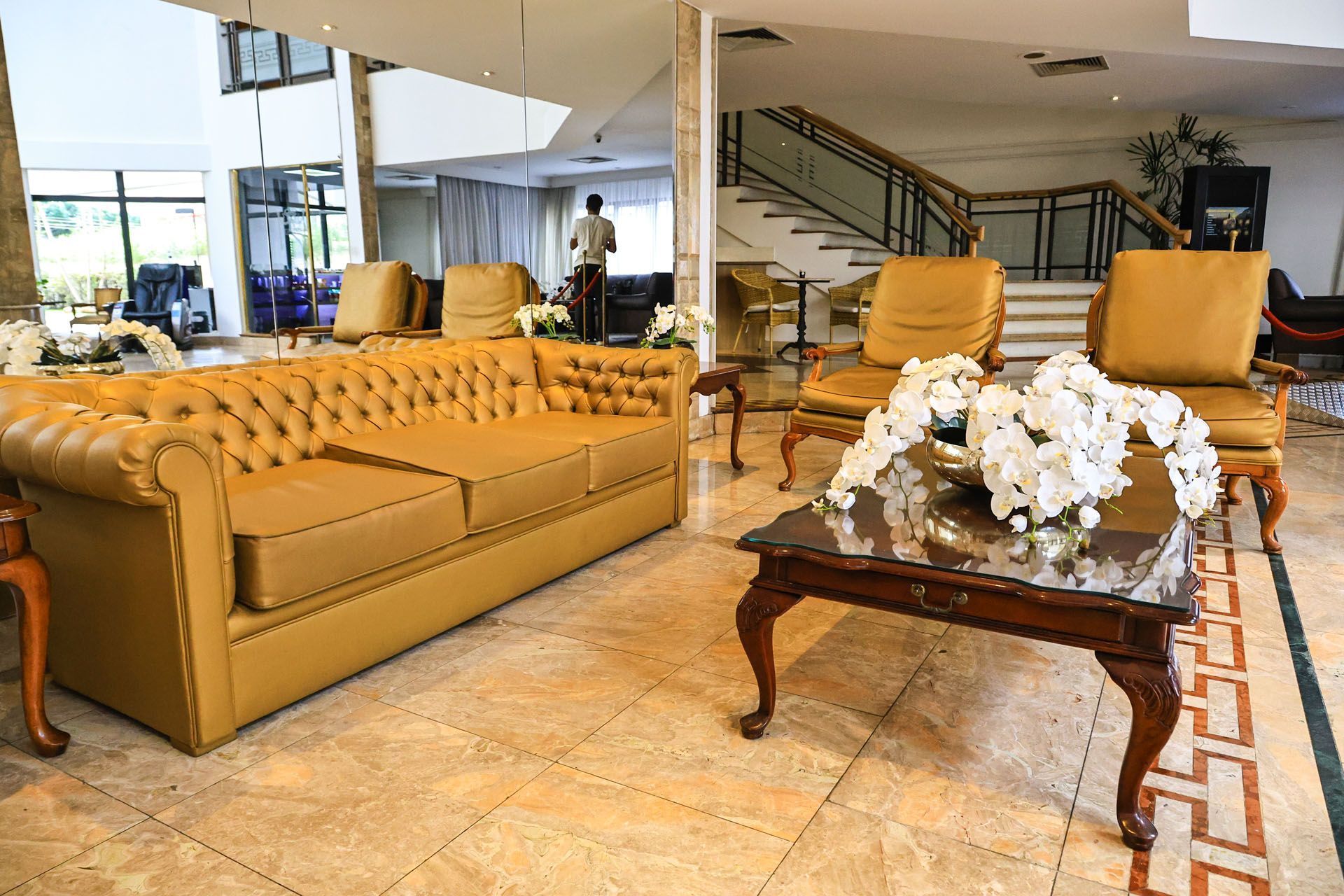 Amplo saguão de hotel com sofás bege, mesa de centro de vidro e escada ao fundo.