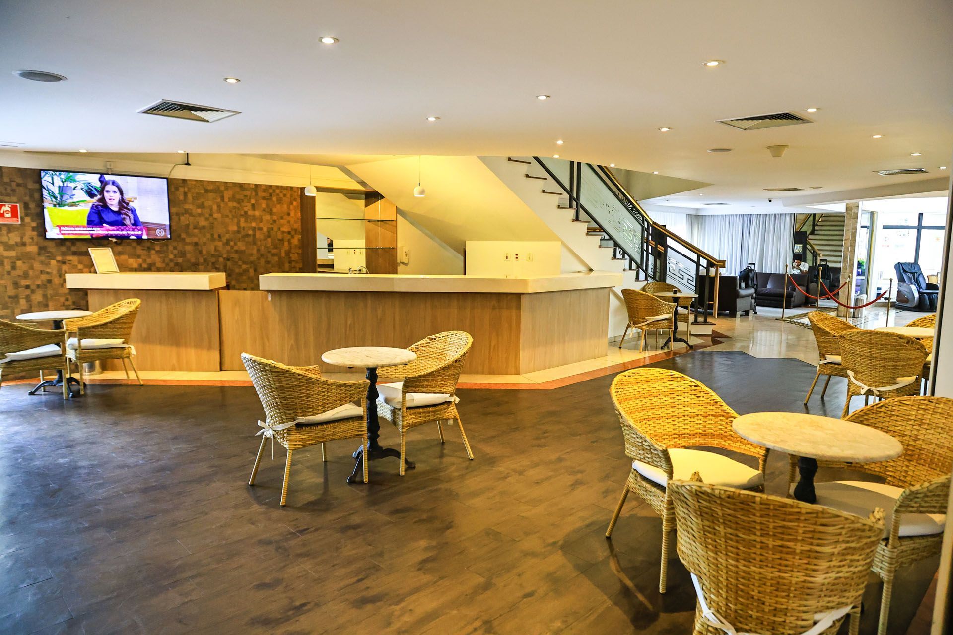 Bar moderno no lobby do hotel com cadeiras de vime, mesas redondas, escadaria e TV em iluminação aconchegante.