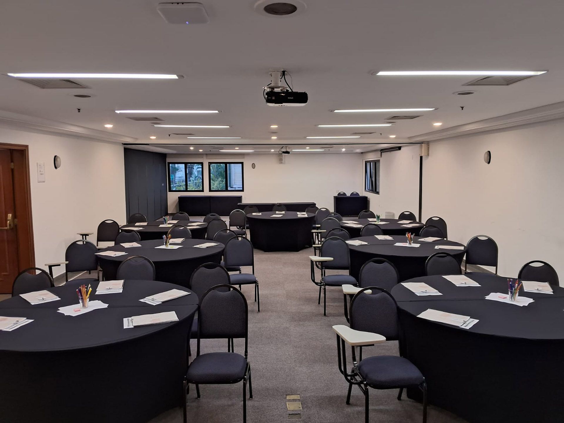 Sala de conferências vazia com fileiras de mesas e cadeiras cobertas de branco, carpete cinza e cortinas fechadas.