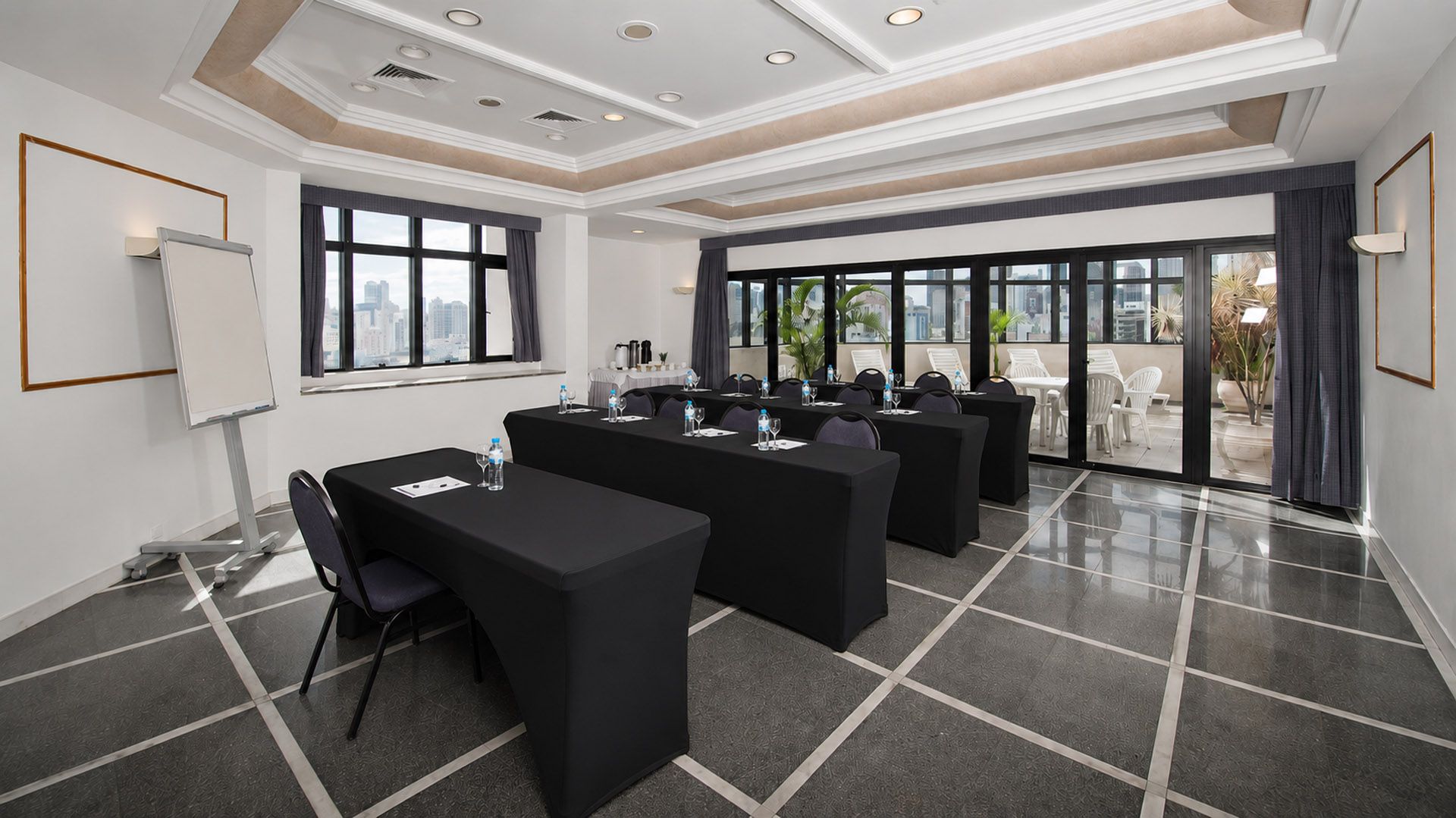 Sala de conferências bem iluminada, preparada para uma reunião, com toalhas de mesa brancas, cadeiras, um flip chart e janelas amplas.