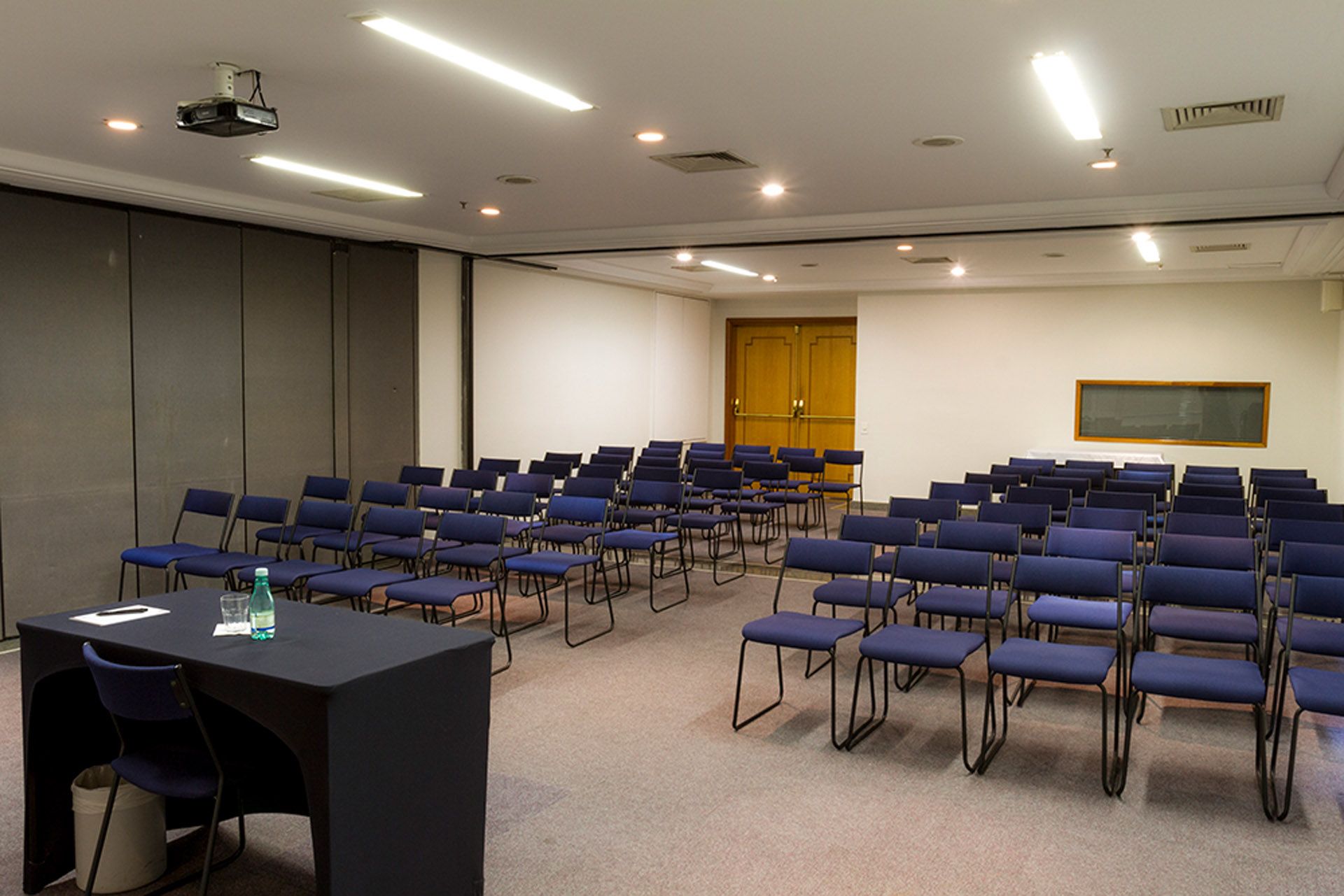 Sala de conferências vazia com fileiras de cadeiras pretas voltadas para uma tela de apresentação.