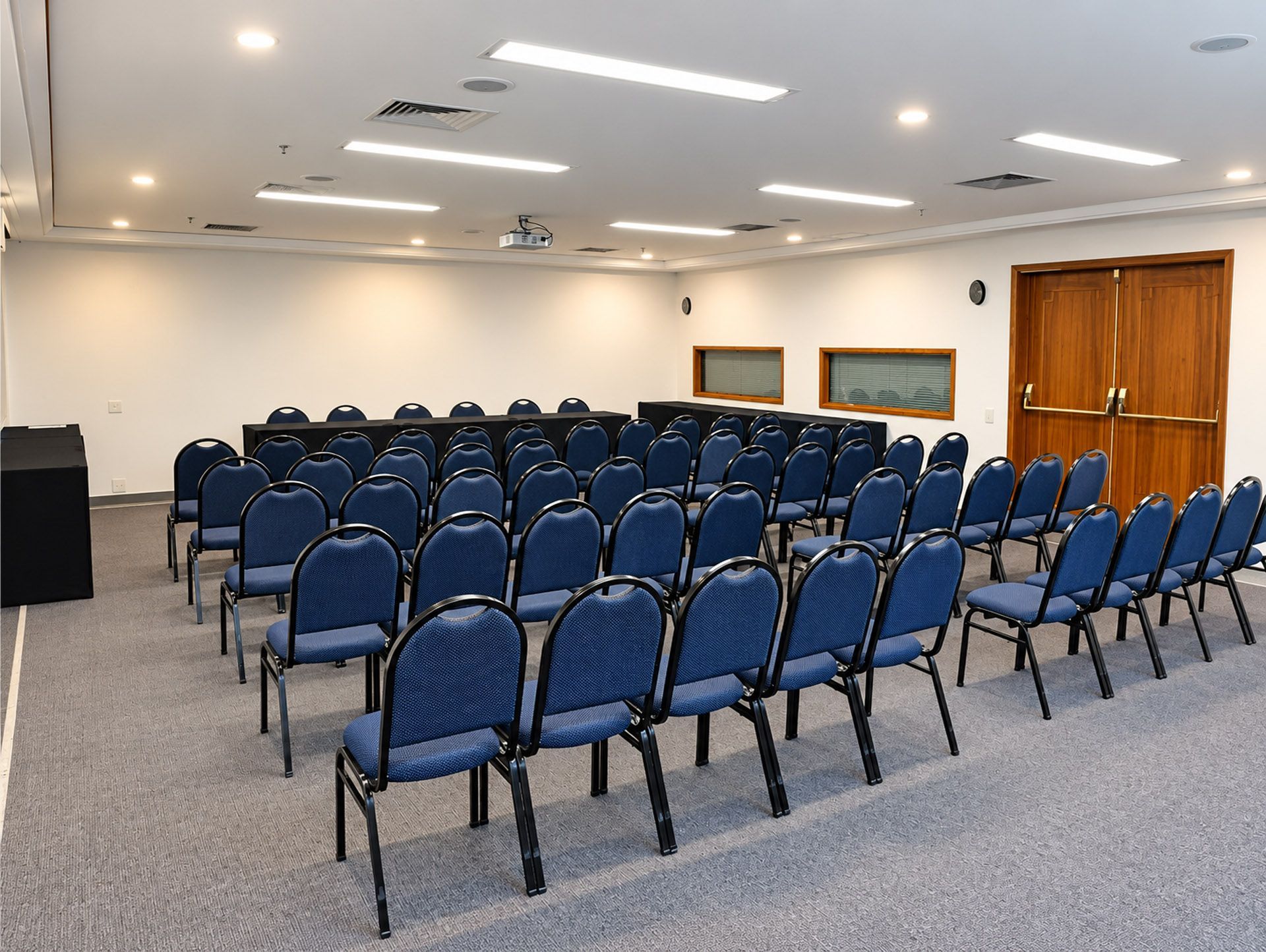 Sala de conferências vazia com fileiras de mesas cobertas de branco e cadeiras pretas sob luzes fortes.