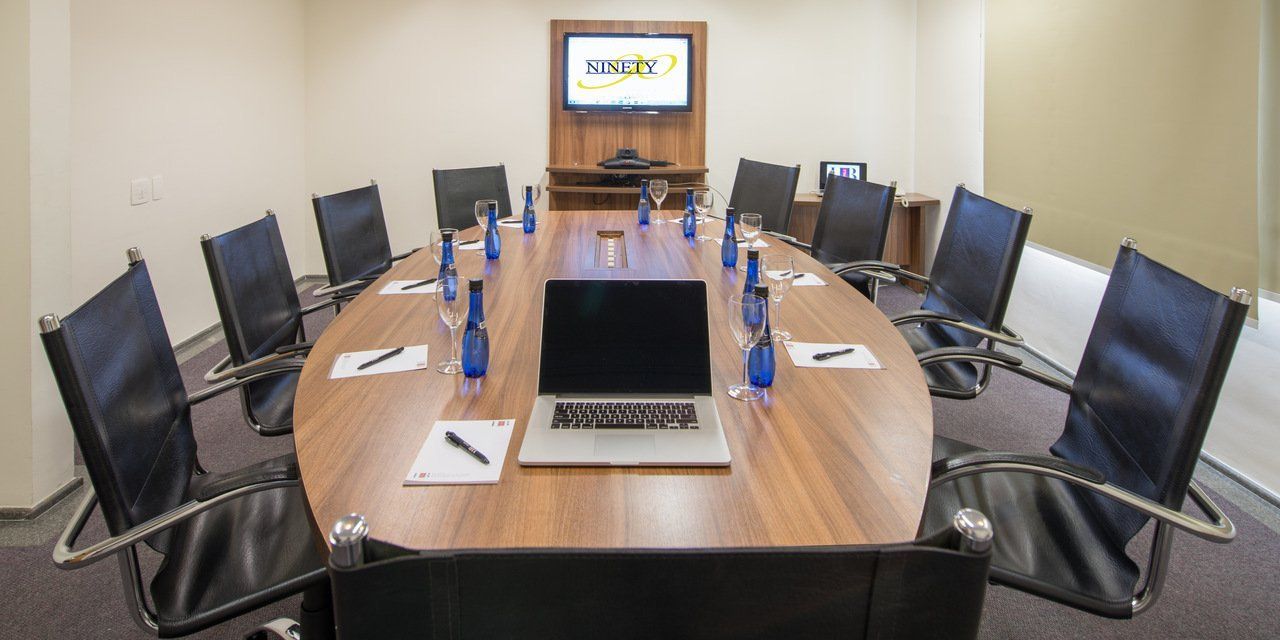 Sala de conferências com uma mesa comprida, laptops, garrafas de água e muitas cadeiras ao redor de uma tela fixada na parede.