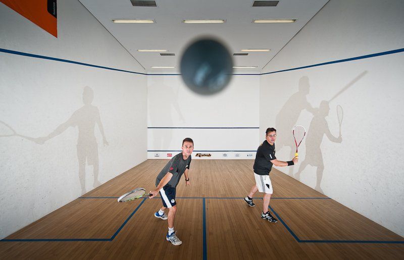 Dois jogadores de squash em uma quadra, se lançando para acertar uma bola perto da parede frontal.