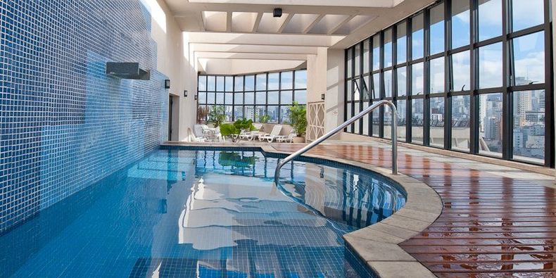 Piscina coberta com águas azuis em formato de curva, paredes revestidas com azulejos e janelas do chão ao teto com vista para um pátio.