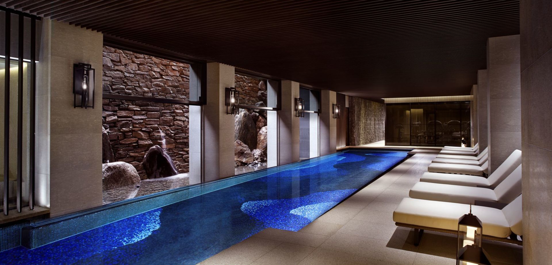 Spa moderno em ambiente fechado com uma longa piscina iluminada em azul, espreguiçadeiras e paredes de pedra texturizada.