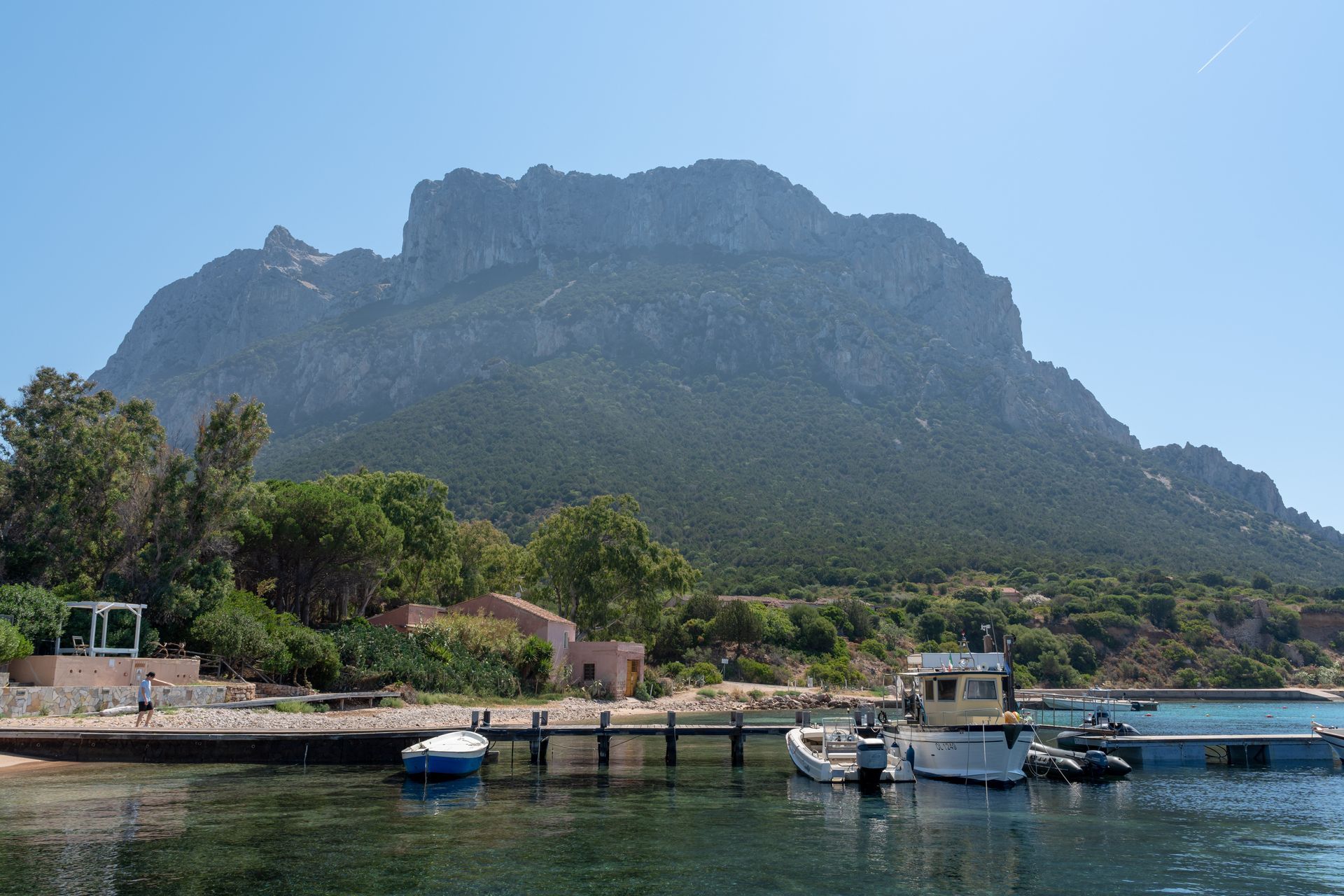 Cinema all'aperto sulla spiaggia; sedie verdi vuote di fronte a uno schermo bianco. Montagne sullo sfondo, giornata di sole.