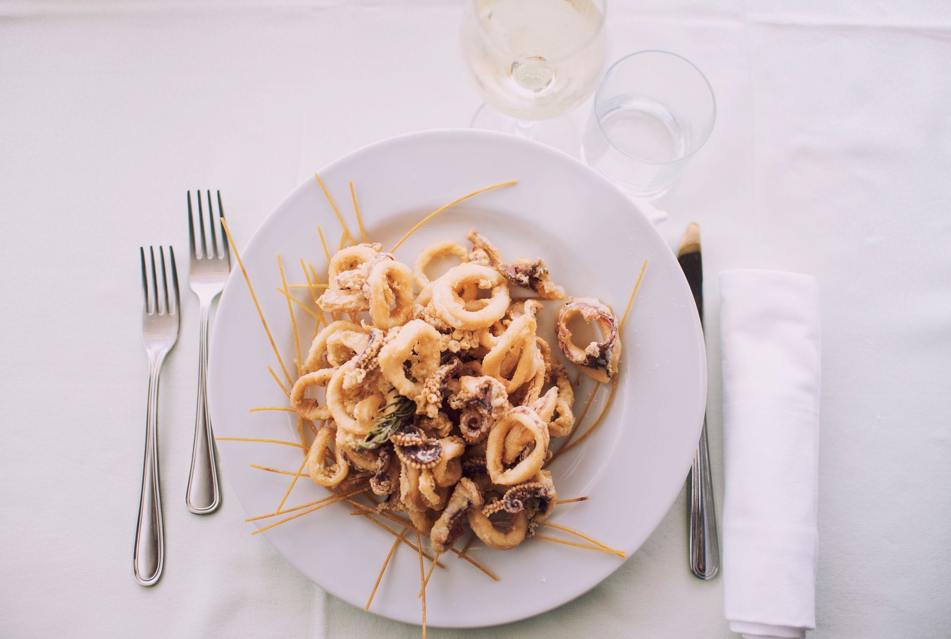 Piatto di calamari fritti con spaghetti su un tavolo bianco, apparecchiato con posate, un tovagliolo e due bicchieri.