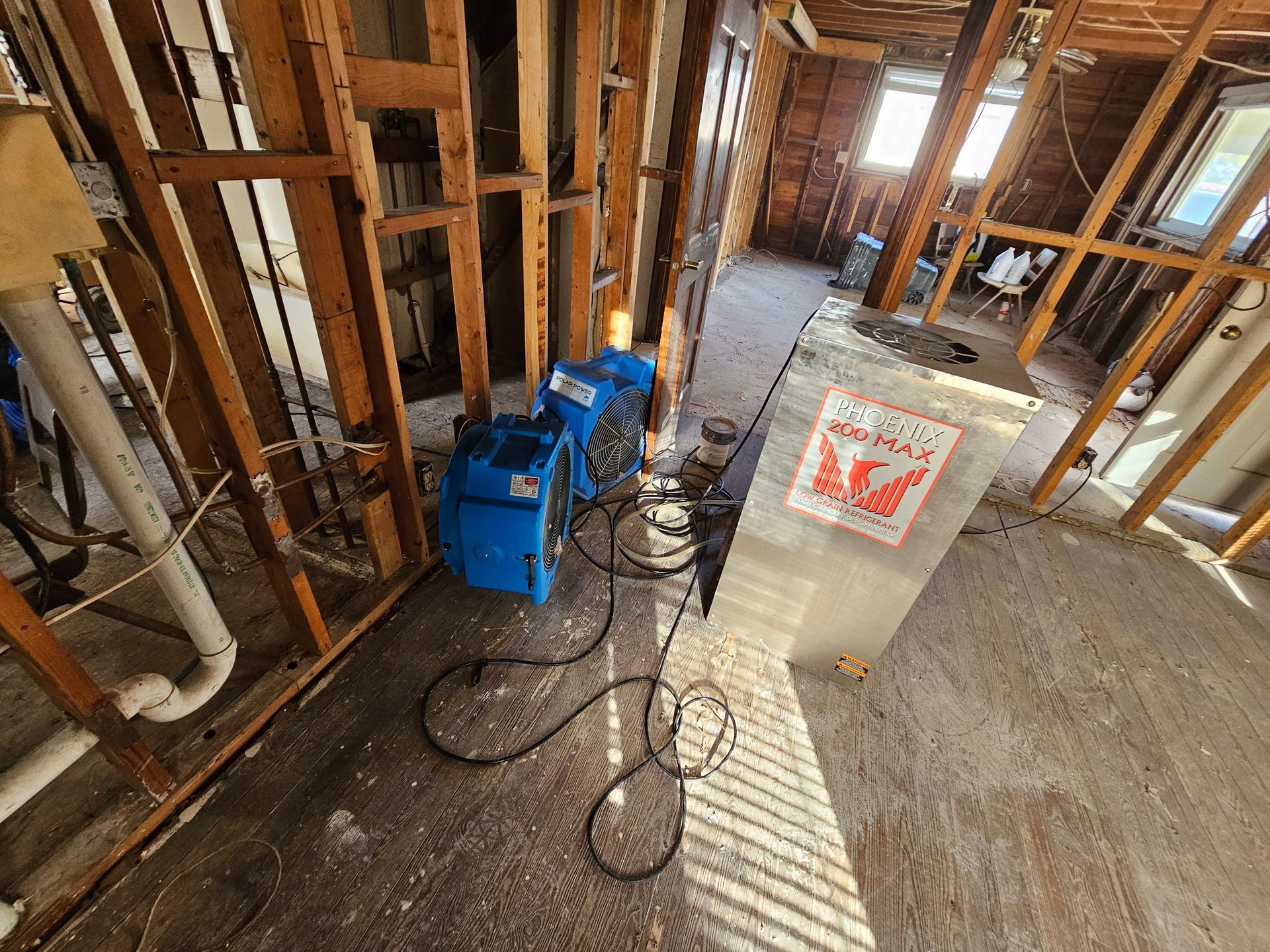 Interior view of a house undergoing renovation. Exposed wooden framing, blue air movers, and debris on the floor.
