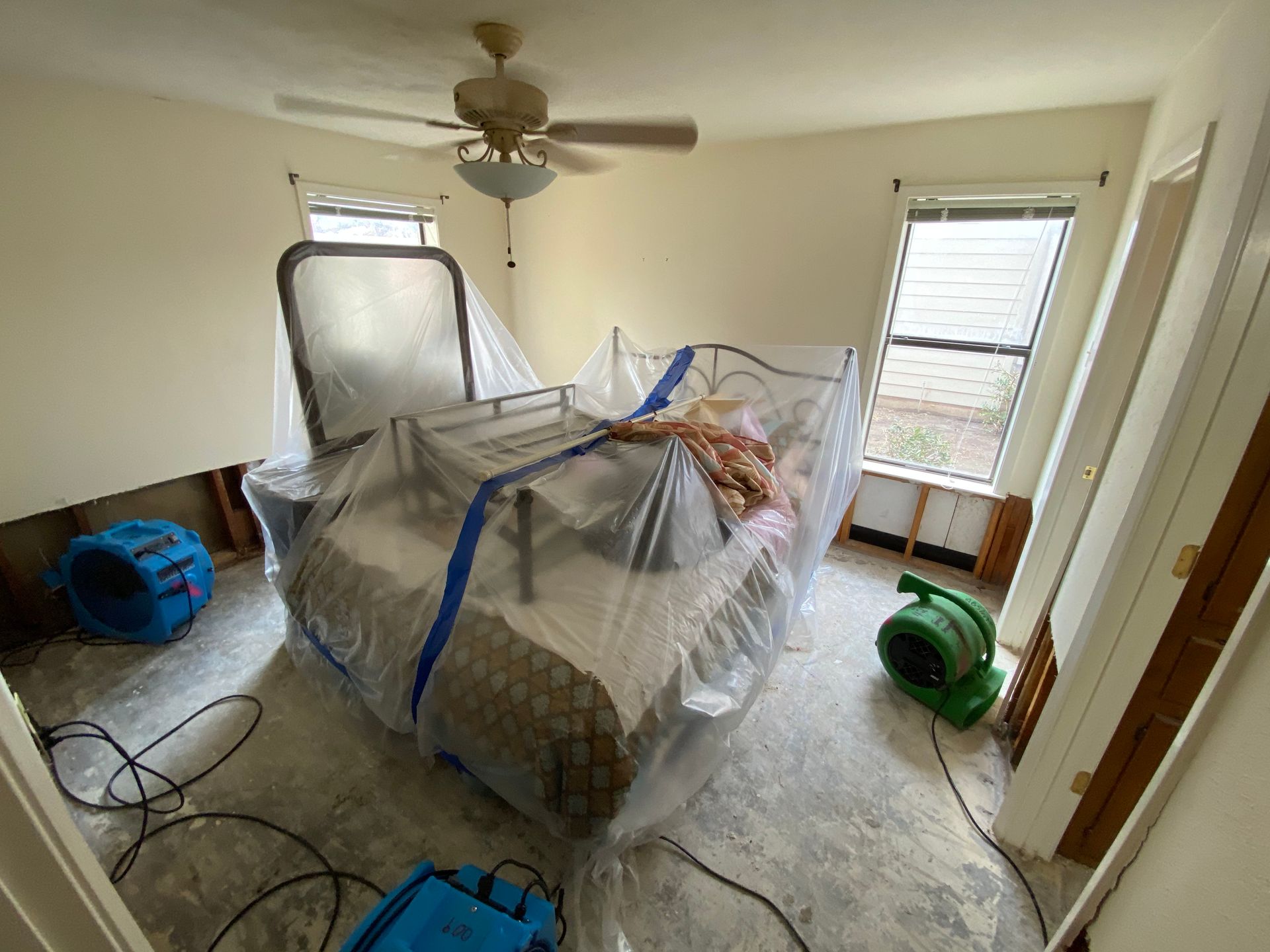 Bedroom with water damage, covered bed, dehumidifiers, and open doorway.