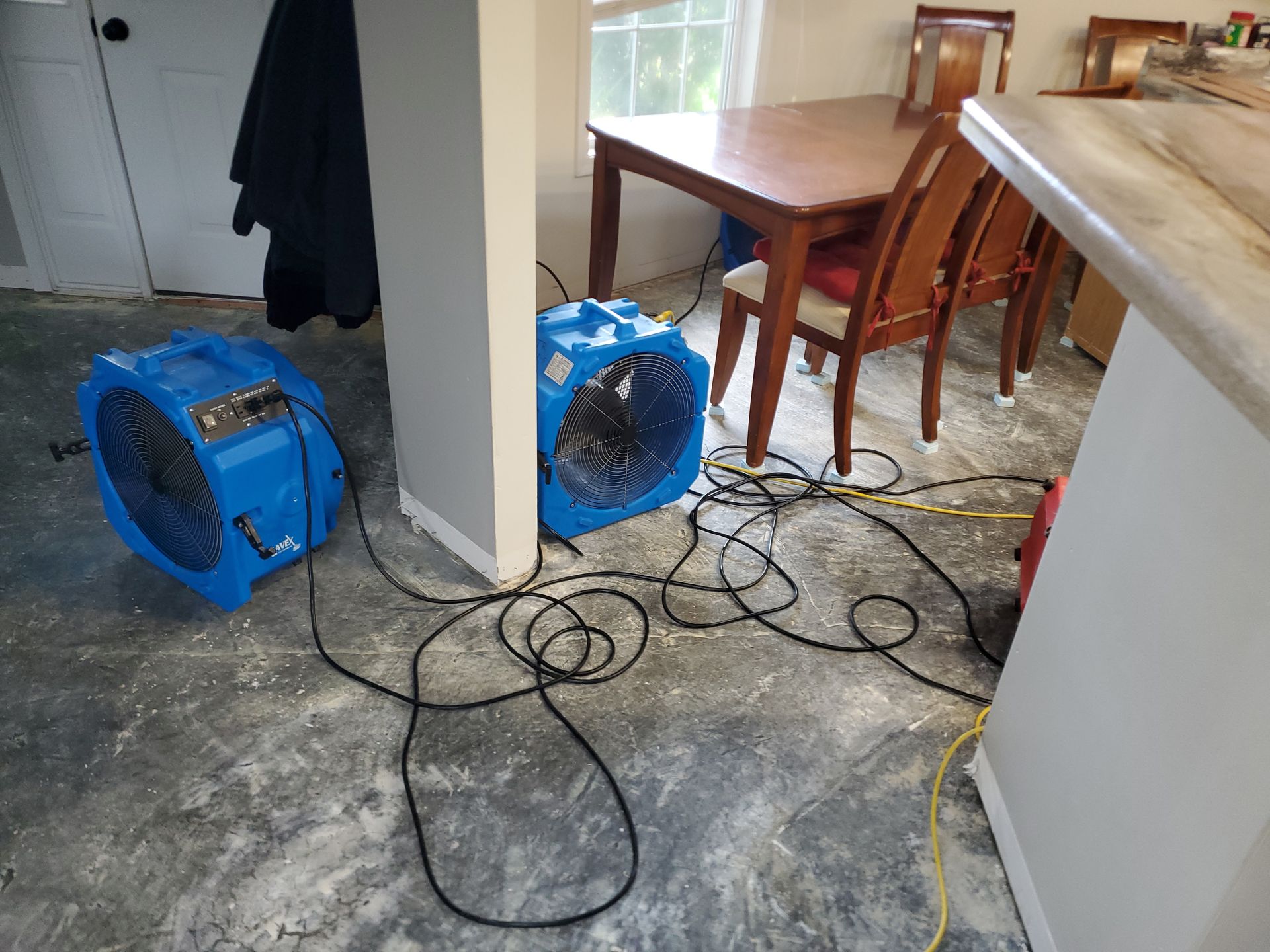 Two blue fans with tangled cords on a concrete floor, near a dining table in a room.
