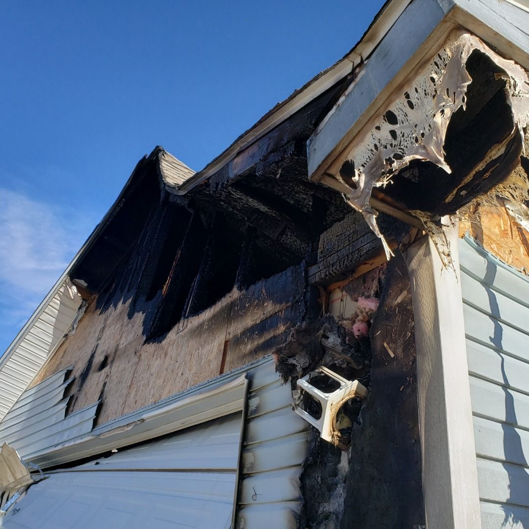 Exterior view of a house showing severe fire damage; charred roof, siding, and exposed structure.