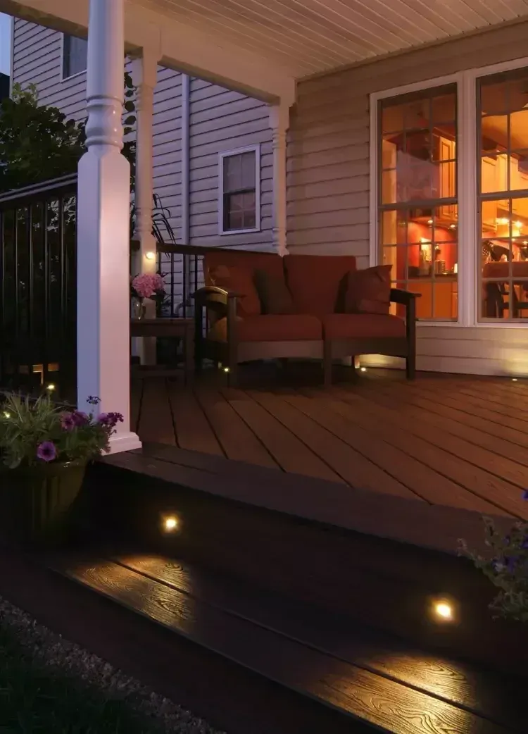 An illuminated wooden deck at dusk, featuring built-in stair lights, a cozy loveseat, and a white porch column.