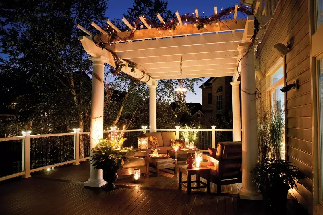 A cozy, illuminated backyard deck at twilight, featuring a pergola, comfortable patio furniture, and warm ambient lighting.
