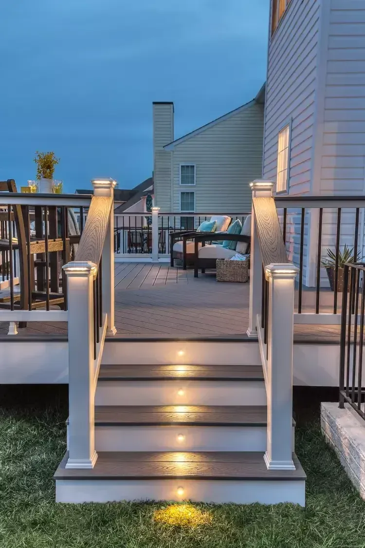 A deck with illuminated stairs leading to a patio area featuring outdoor furniture at twilight.