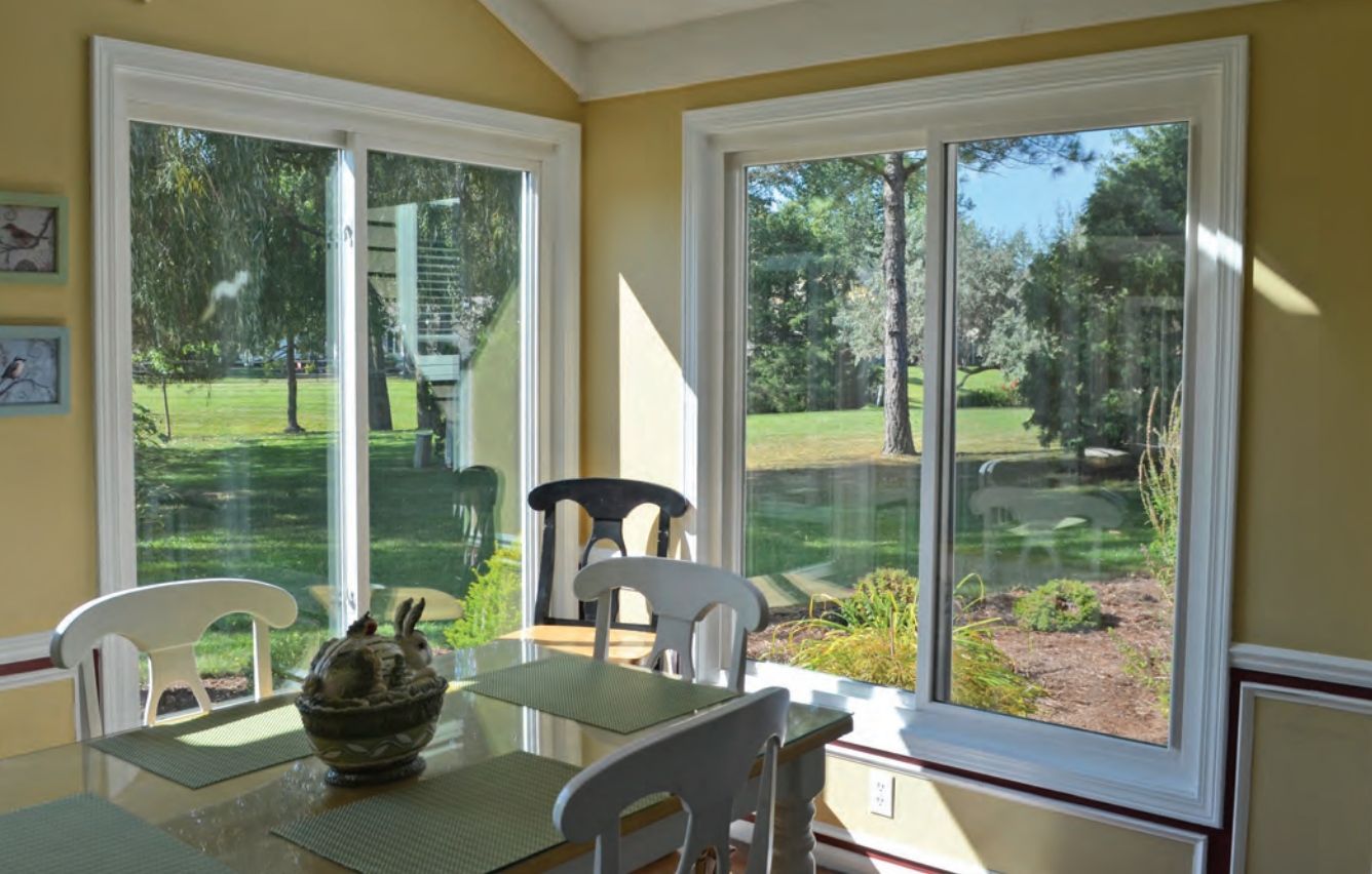 Two wicker armchairs with white cushions sit on a porch in front of a dark-framed double window and hanging greenery.