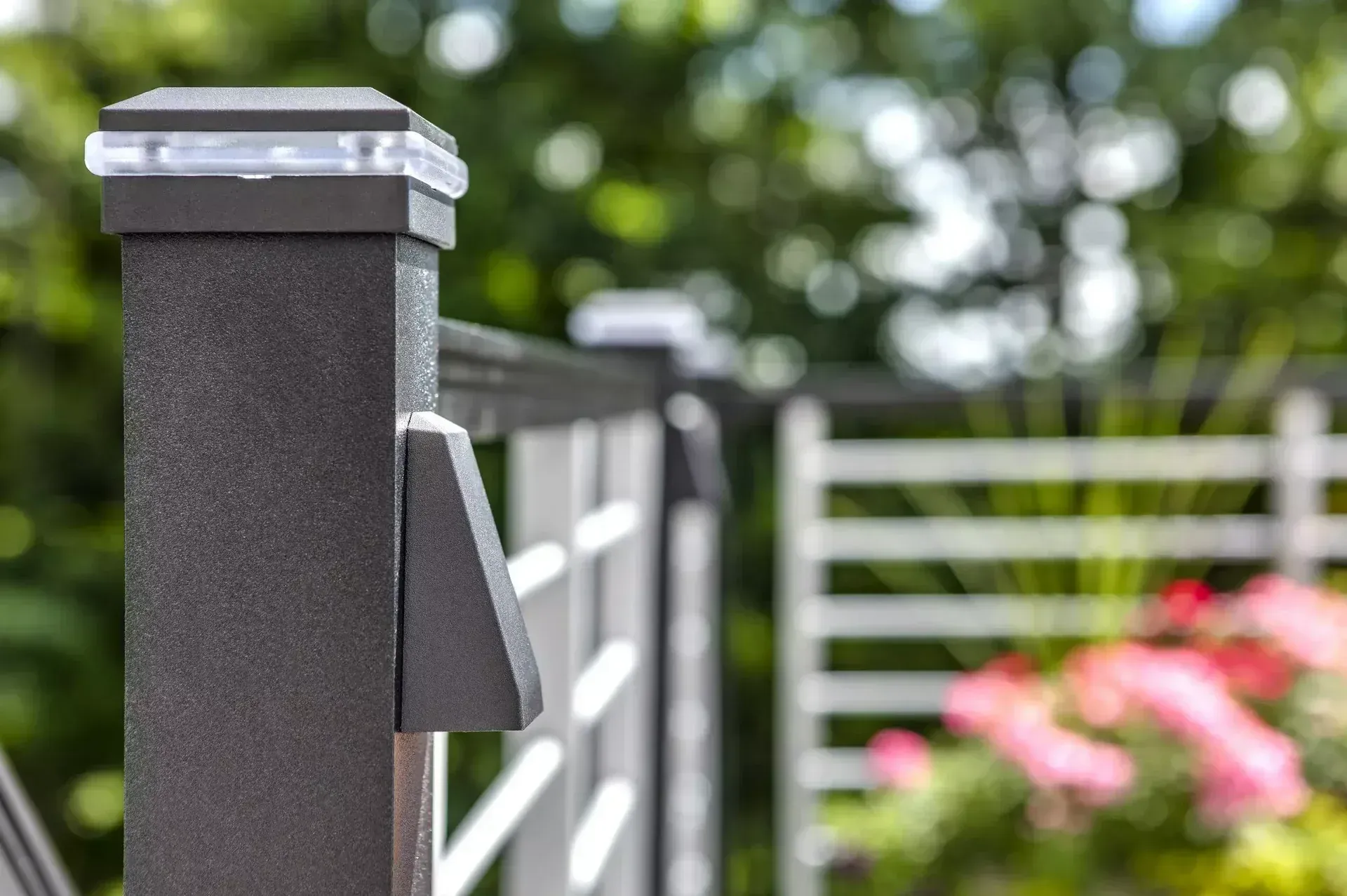 A dark, textured metal fence post with a solar-powered cap light and mounted accent light, set against a blurred garden.