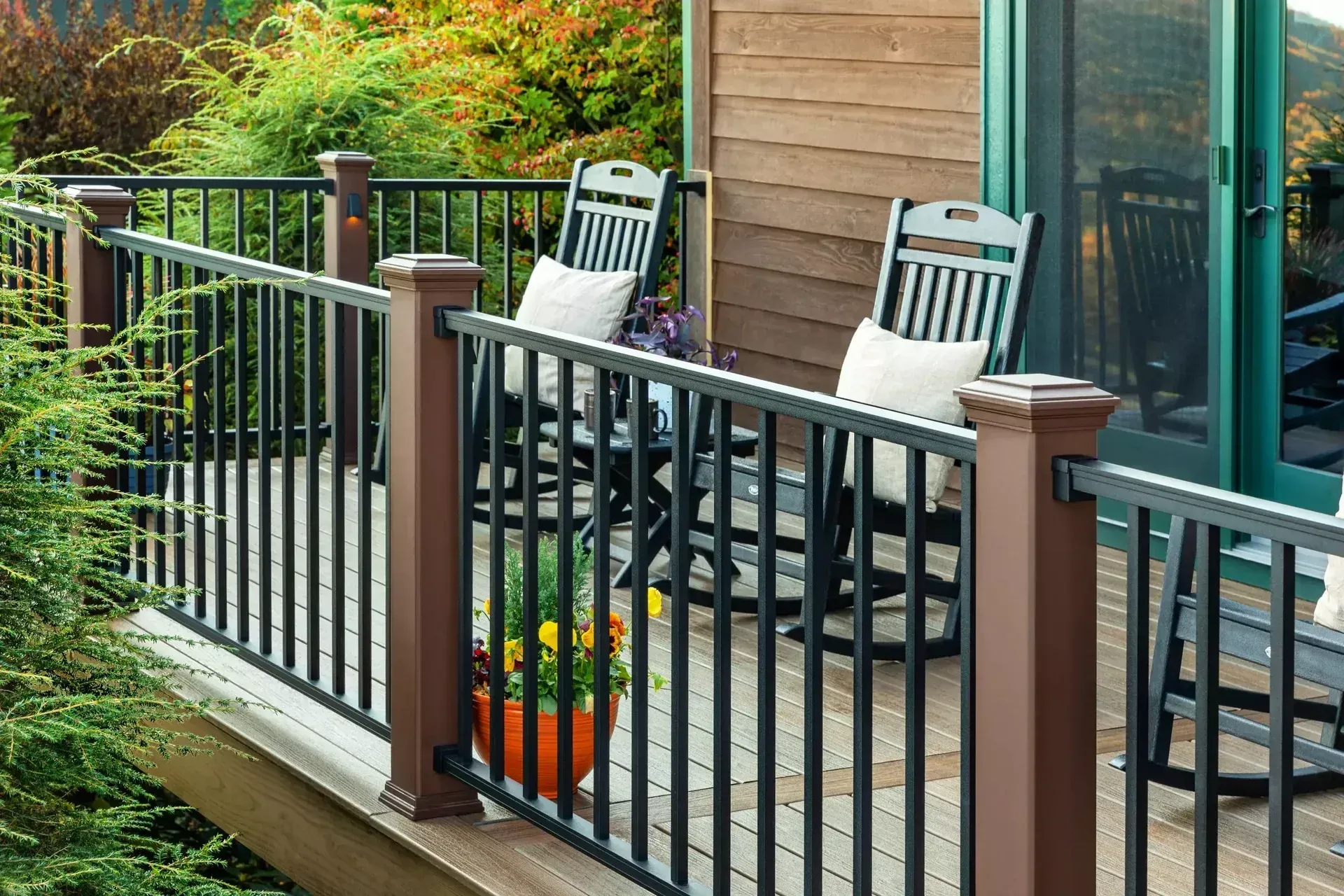 A deck with black railings and two dark wooden rocking chairs with white pillows, set against a background of green trees.
