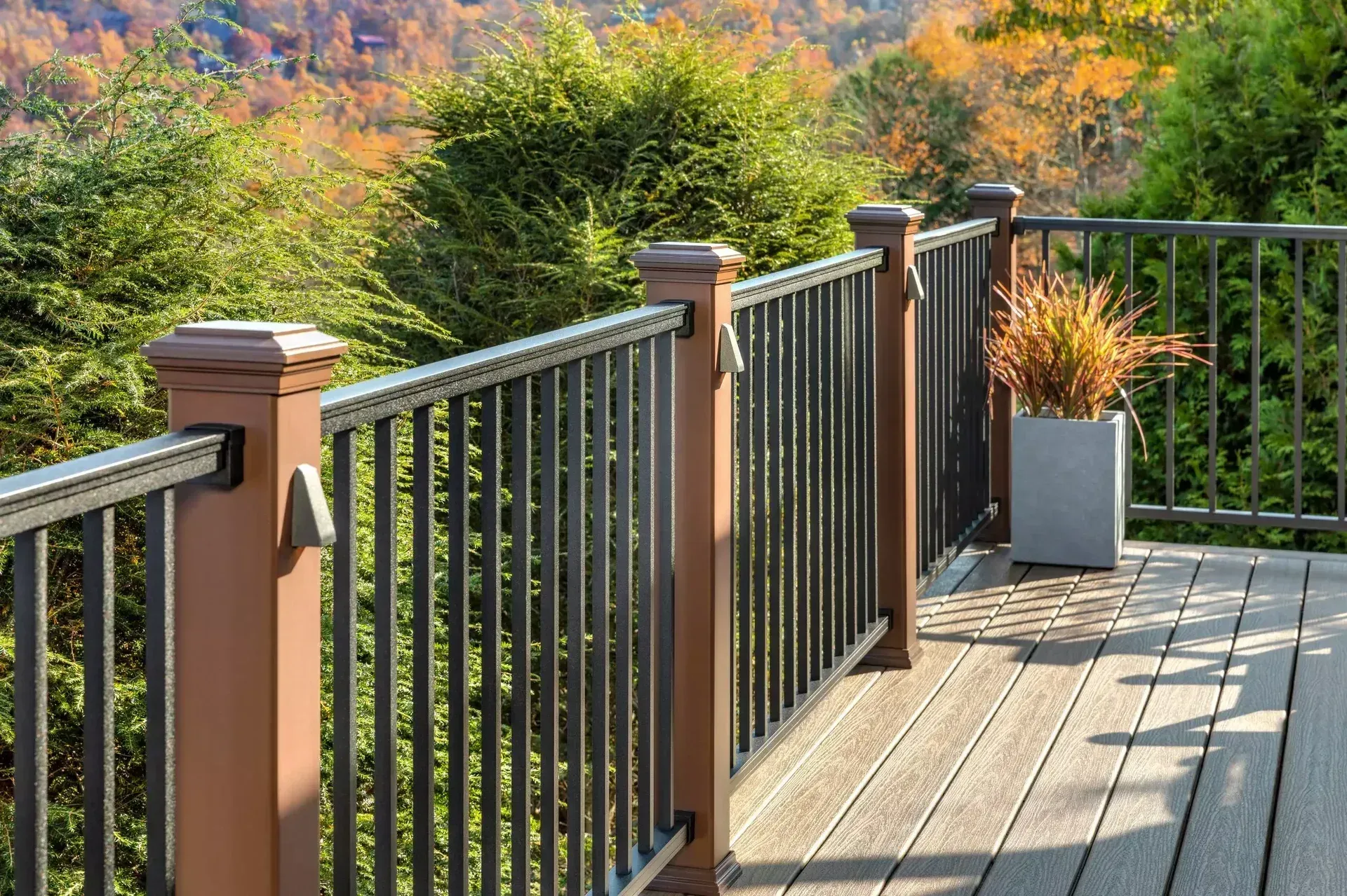 A brown composite deck with dark metal railings overlooking lush green and autumn-colored trees in the background.