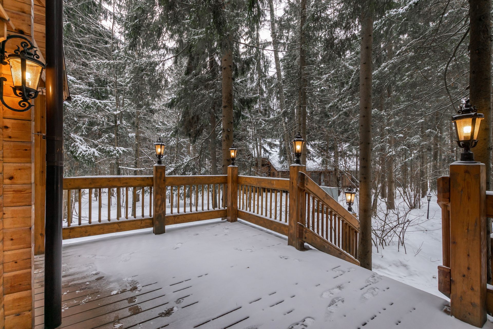 A wooden deck covered in a light dusting of snow, surrounded by snow-dusted pine trees and illuminated by wall sconces.