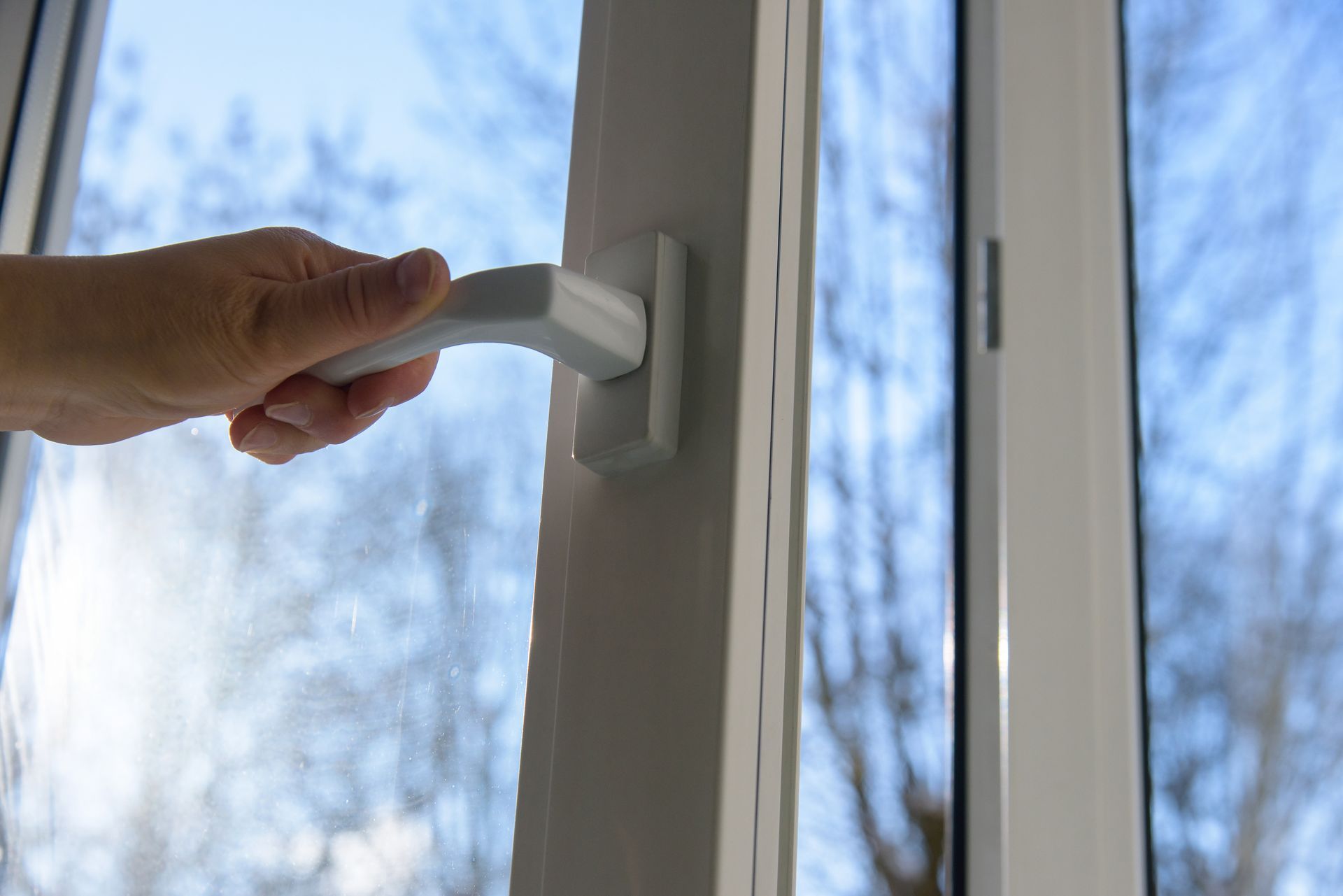 Hand gripping a white window handle beside a bright window with bare trees outside Hand gripping a white window handle beside a bright window with bare trees outside