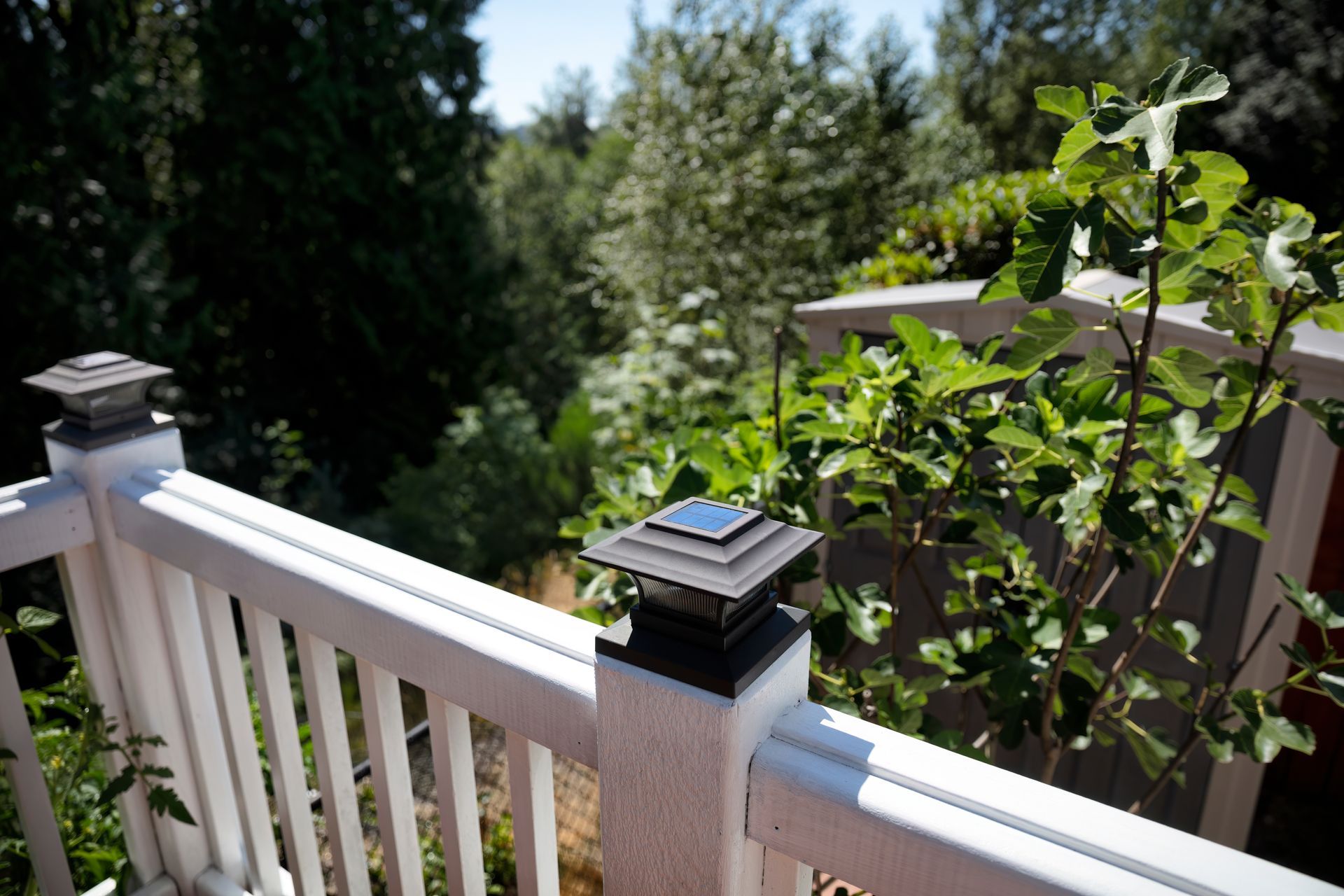 A white deck railing with square solar-powered post cap lights, overlooking a lush green wooded area.