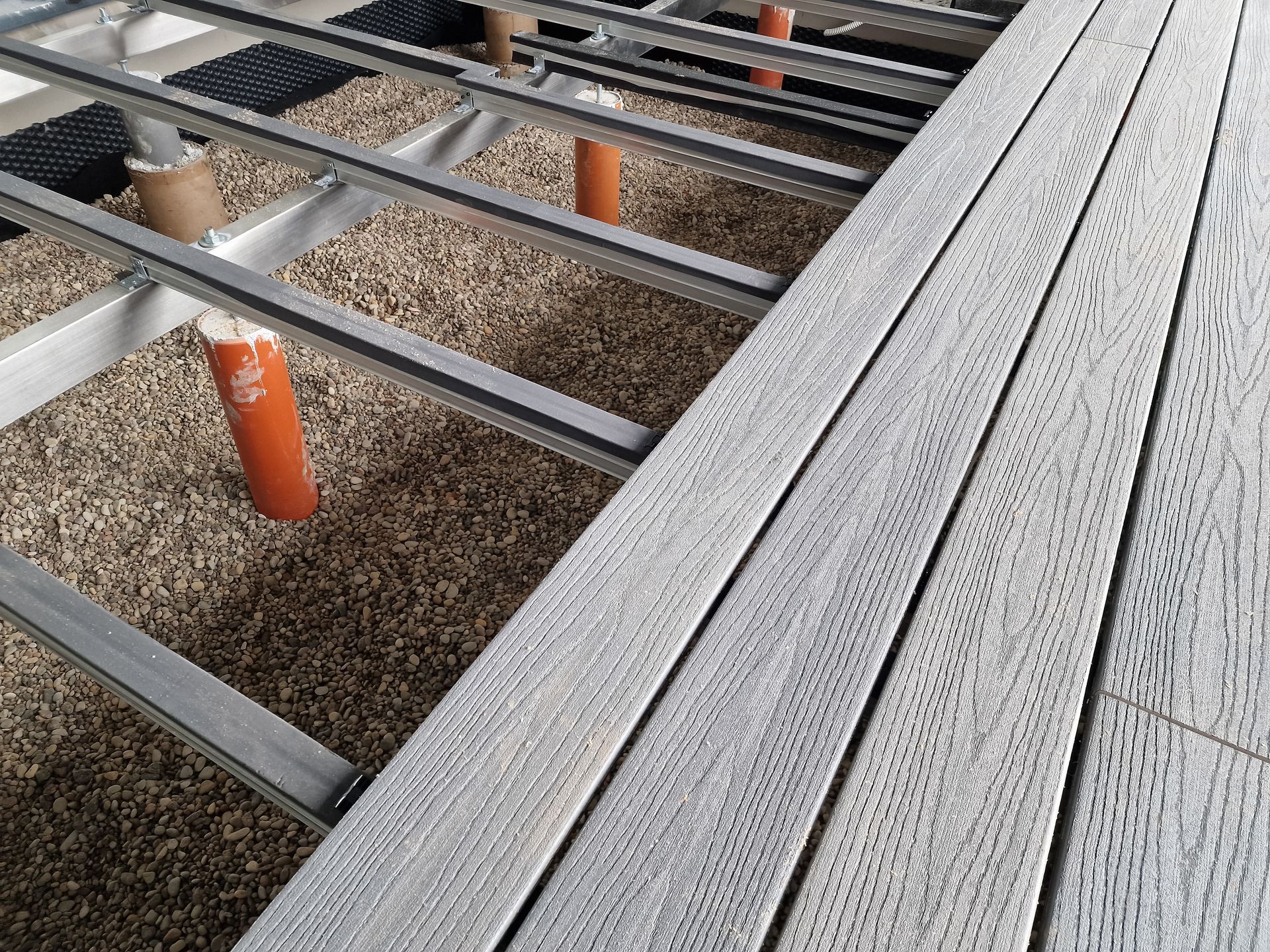 Gray composite deck boards being installed on a metal frame over a gravel base with orange support pillars.