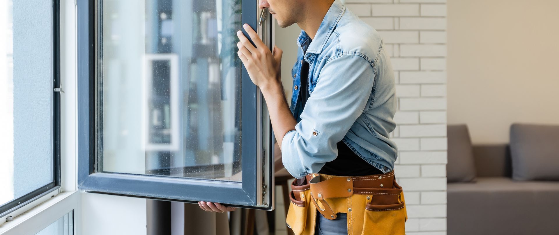 Person in a blue shirt and tool belt fixing a window beside a white brick wall Person in a blue shirt and tool belt fixing a window beside a white brick wall