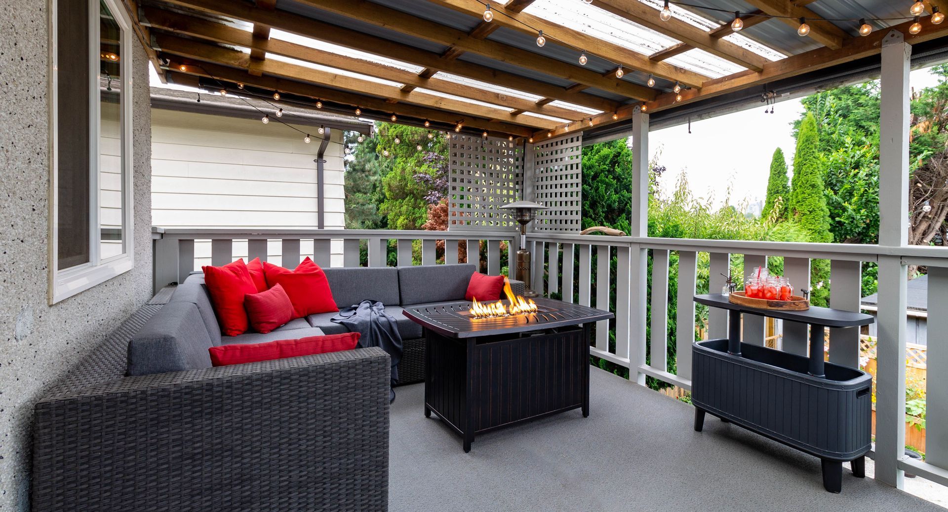 A cozy, covered outdoor patio featuring a gray sectional sofa with red pillows, a square gas fire pit, and string lights.