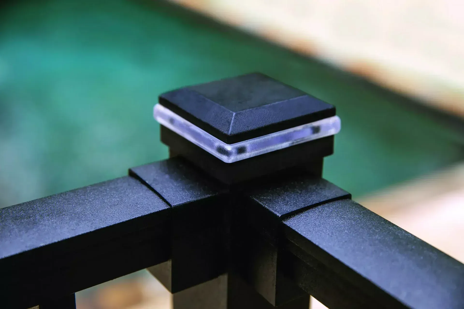 A black, square-topped post cap light with a clear glowing edge sits atop a railing corner, with a blurry green background.