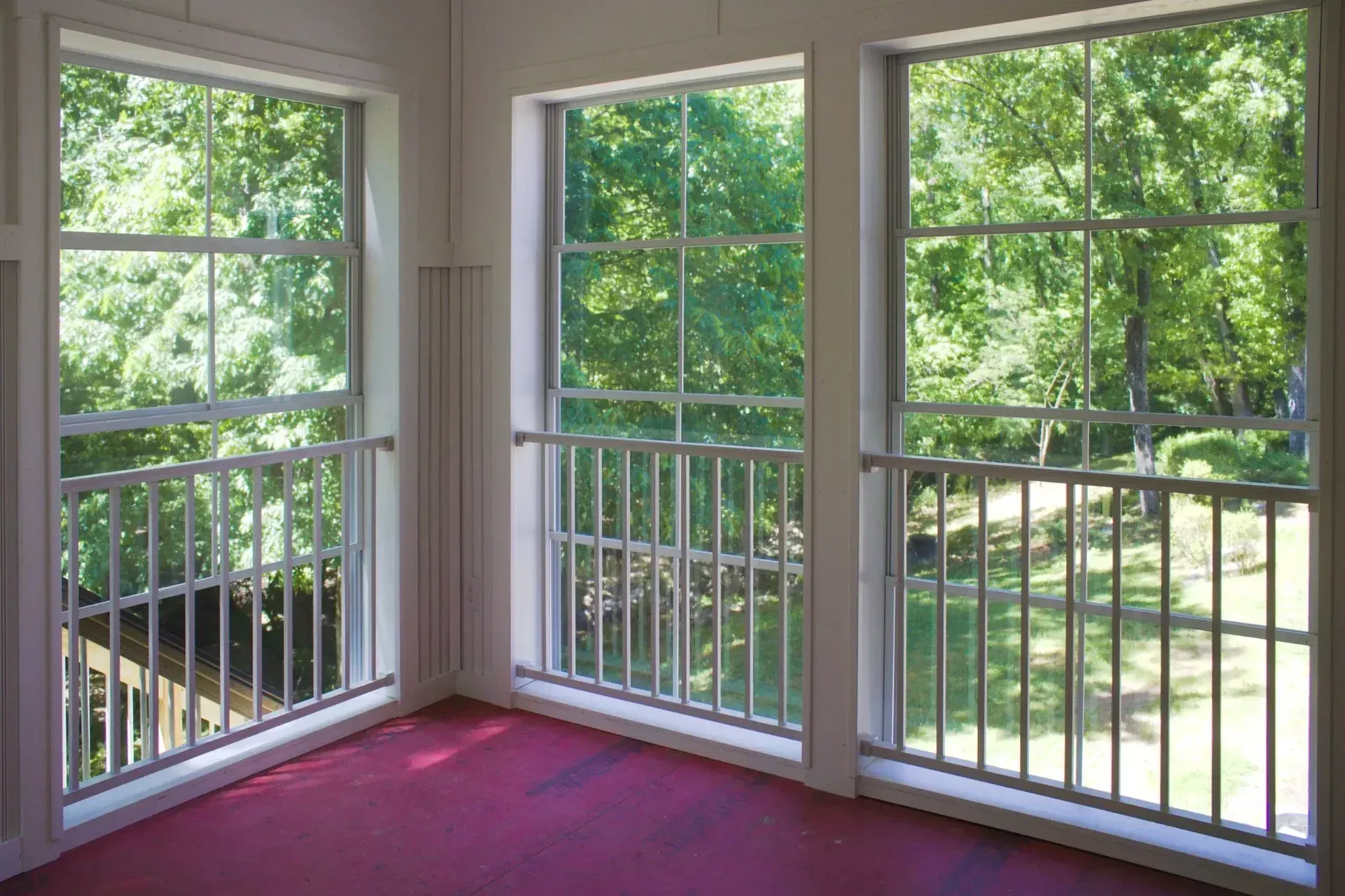 Three large windows with white frames and interior safety railings look out onto a green, tree-filled landscape.