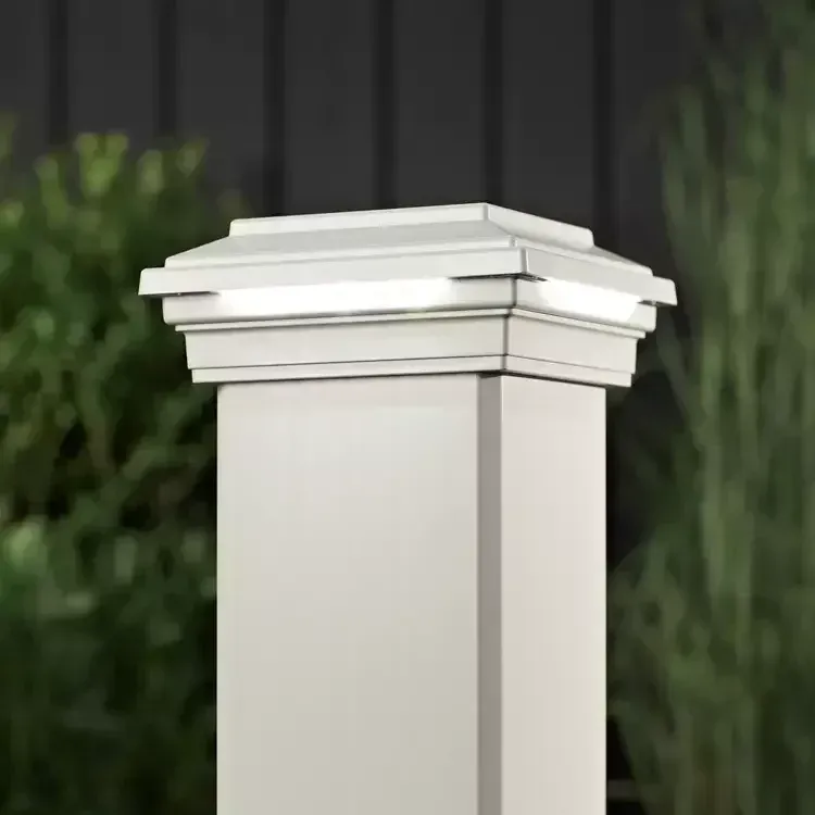 A white, square post cap light glowing on top of a matching fence post, with blurred greenery in the background.