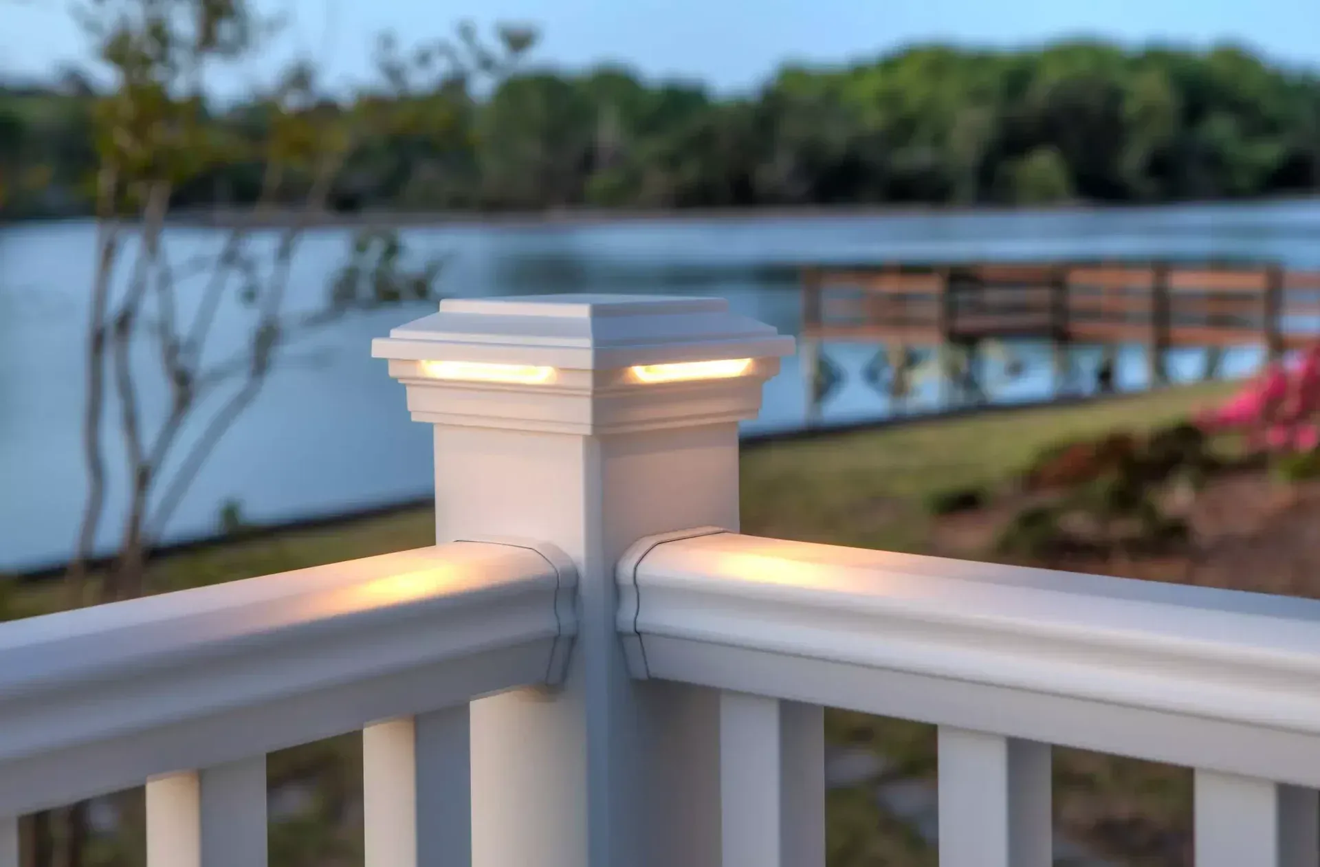 A white deck railing post cap with integrated warm LED accent lighting, overlooking a waterfront scene at dusk.