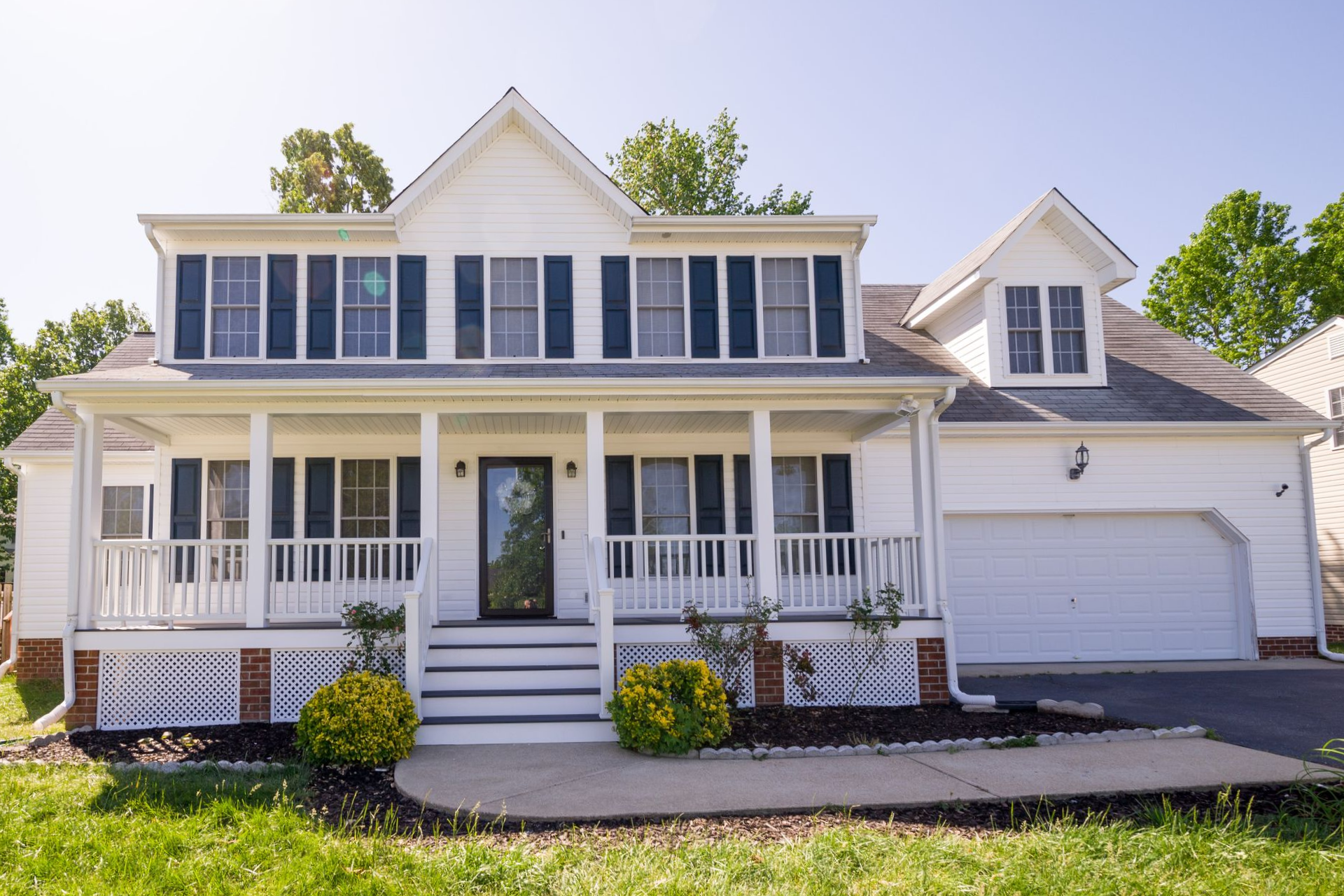 Two-story white suburban house with front porch, dormers, and attached garage in bright daylight