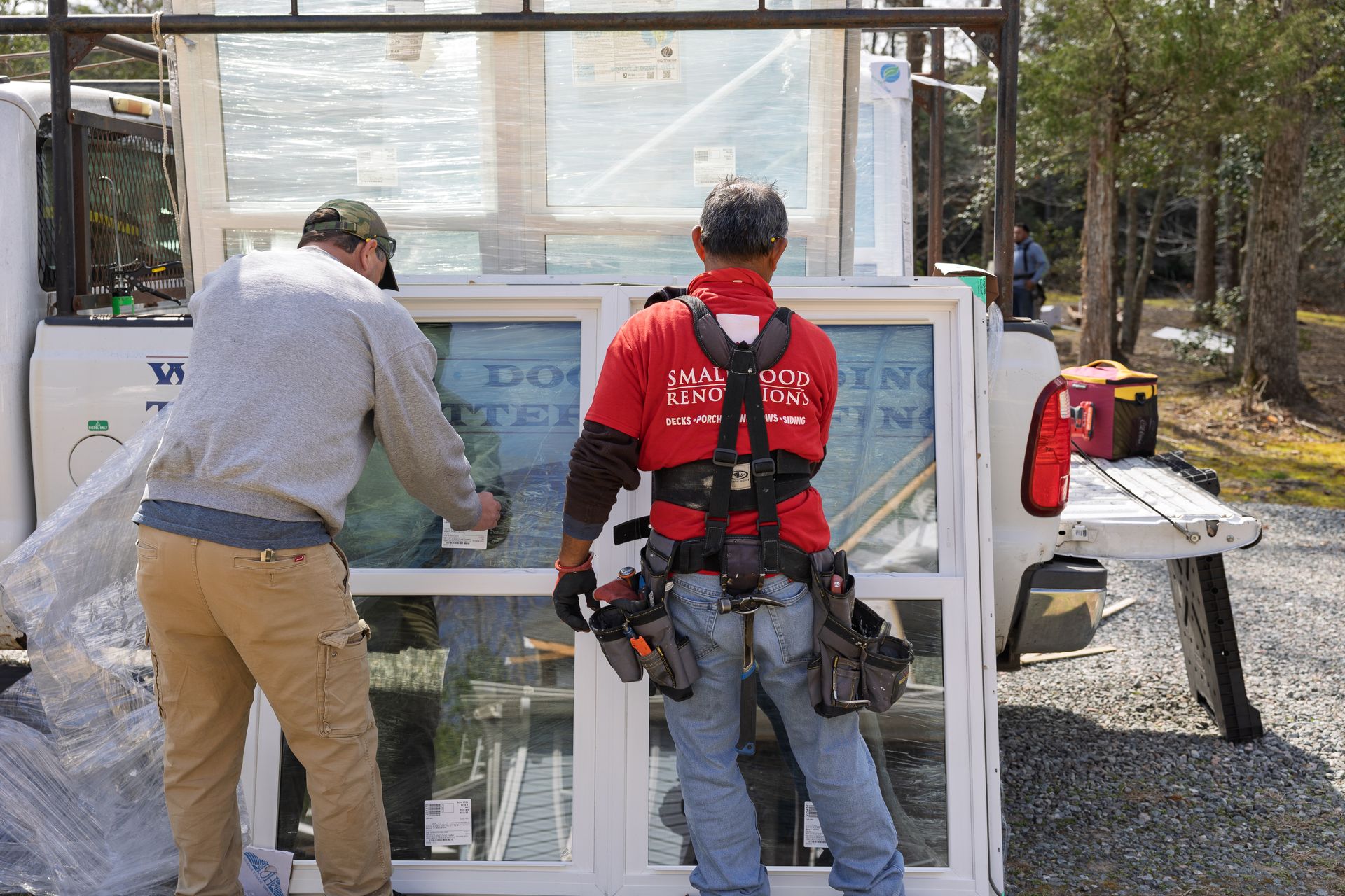 Two workers in a gravel lot unload large glass windows from the back of a utility truck.