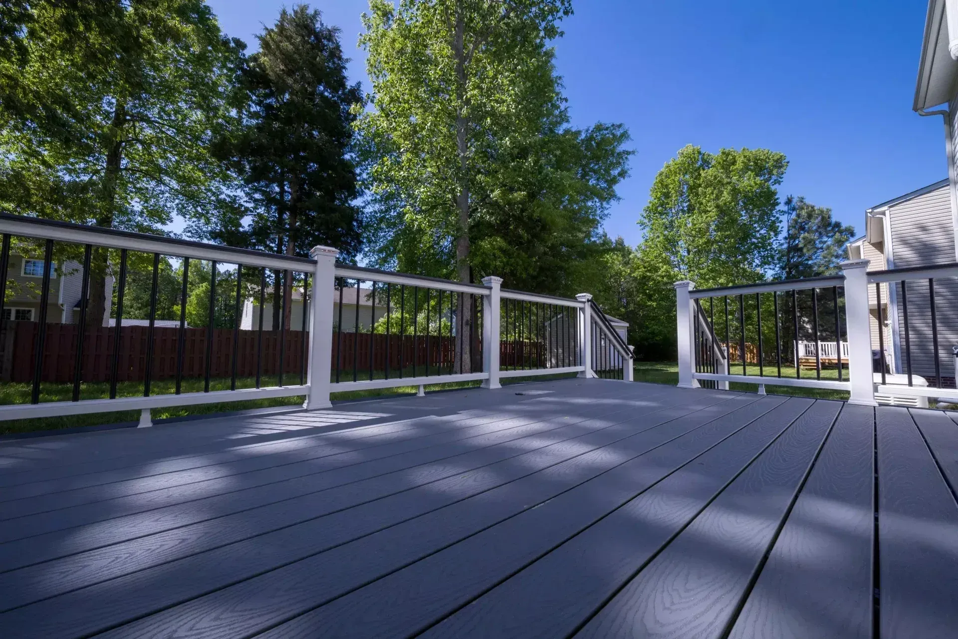 A wide-angle view of a gray composite deck with white railings, looking out toward a backyard with trees and a clear sky.