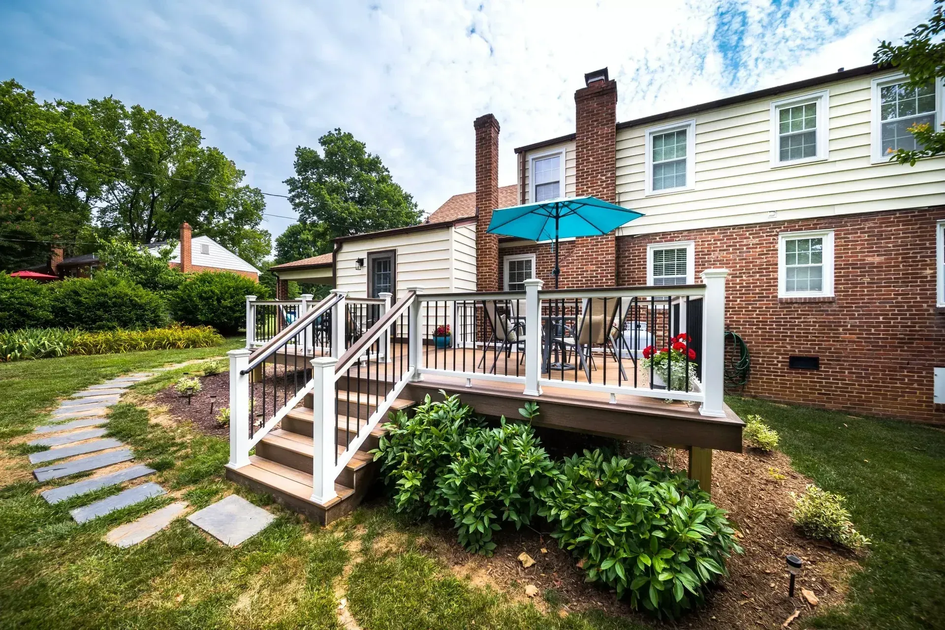 A wooden deck with white railings and a blue umbrella attached to the brick and cream-sided back of a suburban house.