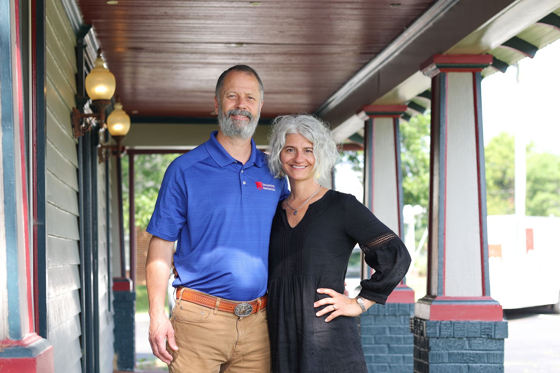Two people standing on a porch, smiling, with a covered roof and red-trimmed columns behind them