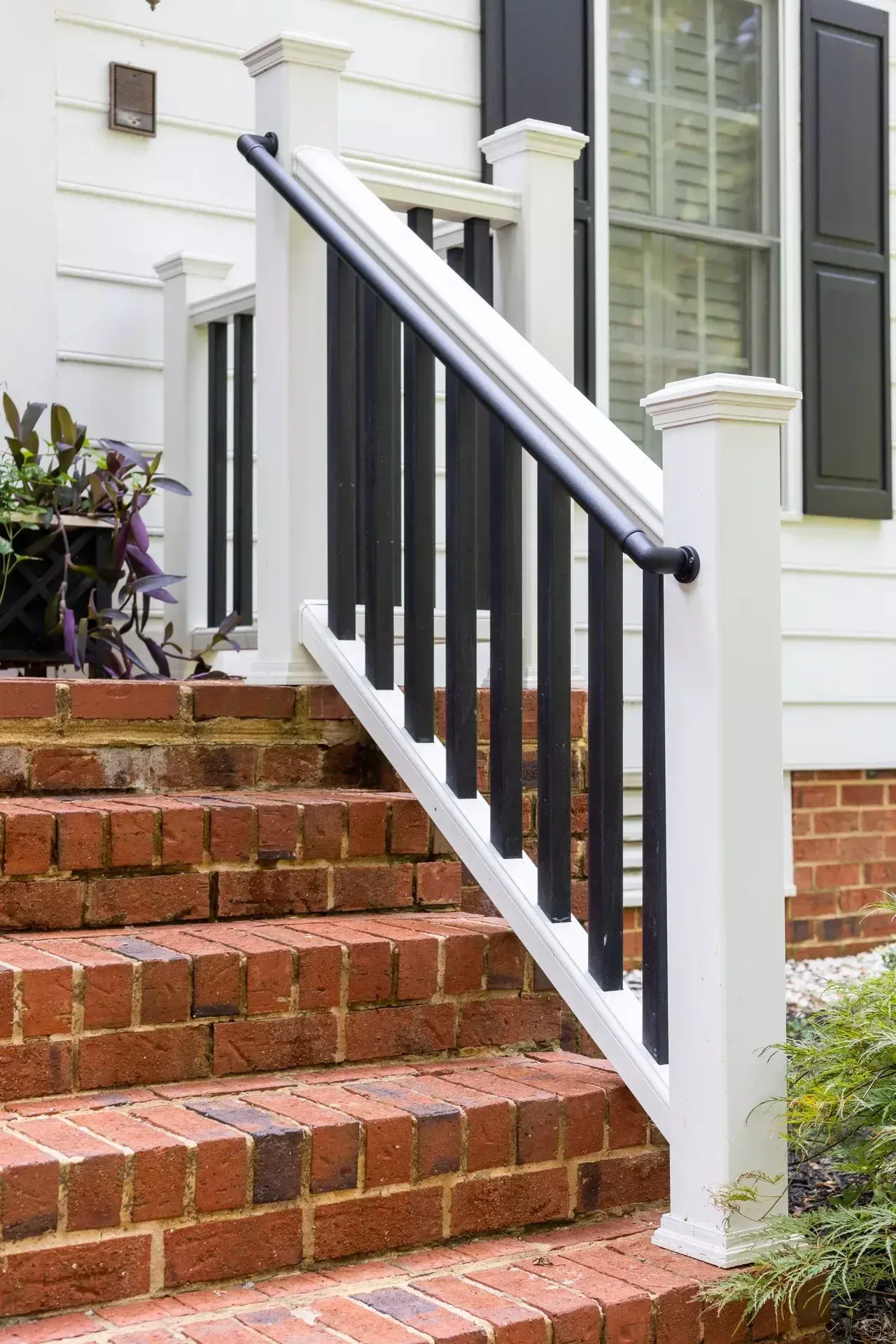 A black metal handrail mounted on white wooden posts along outdoor brick steps leading to a house entrance.