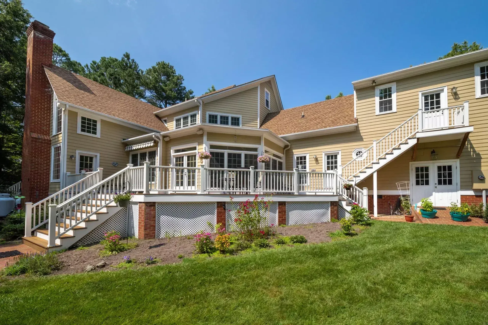 A tan, multi-story house with a wooden deck, a staircase, and a large backyard lawn under a clear blue sky.